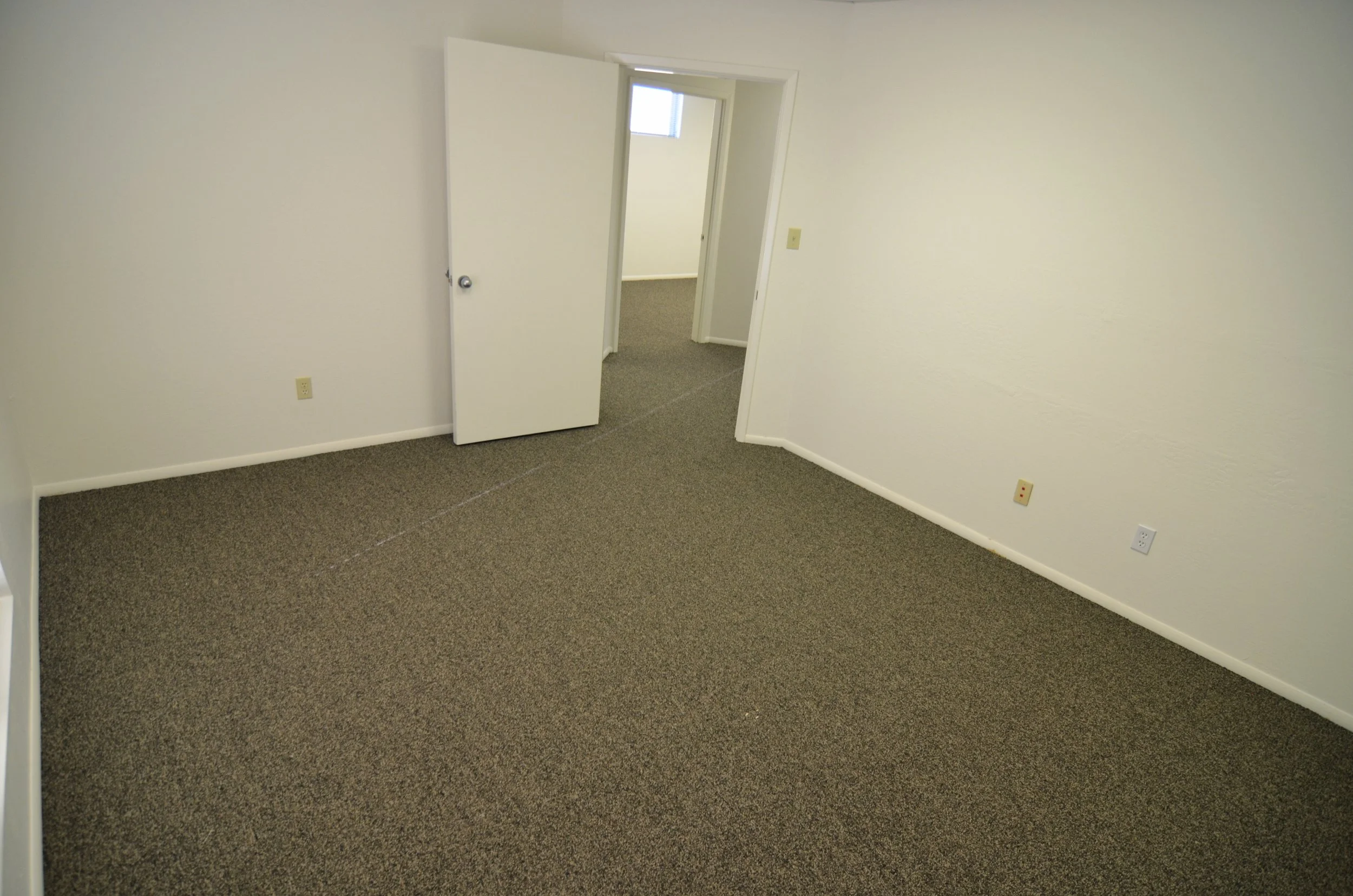 Empty room with beige carpet, white walls, open door leading to another room, and visible electrical outlets.