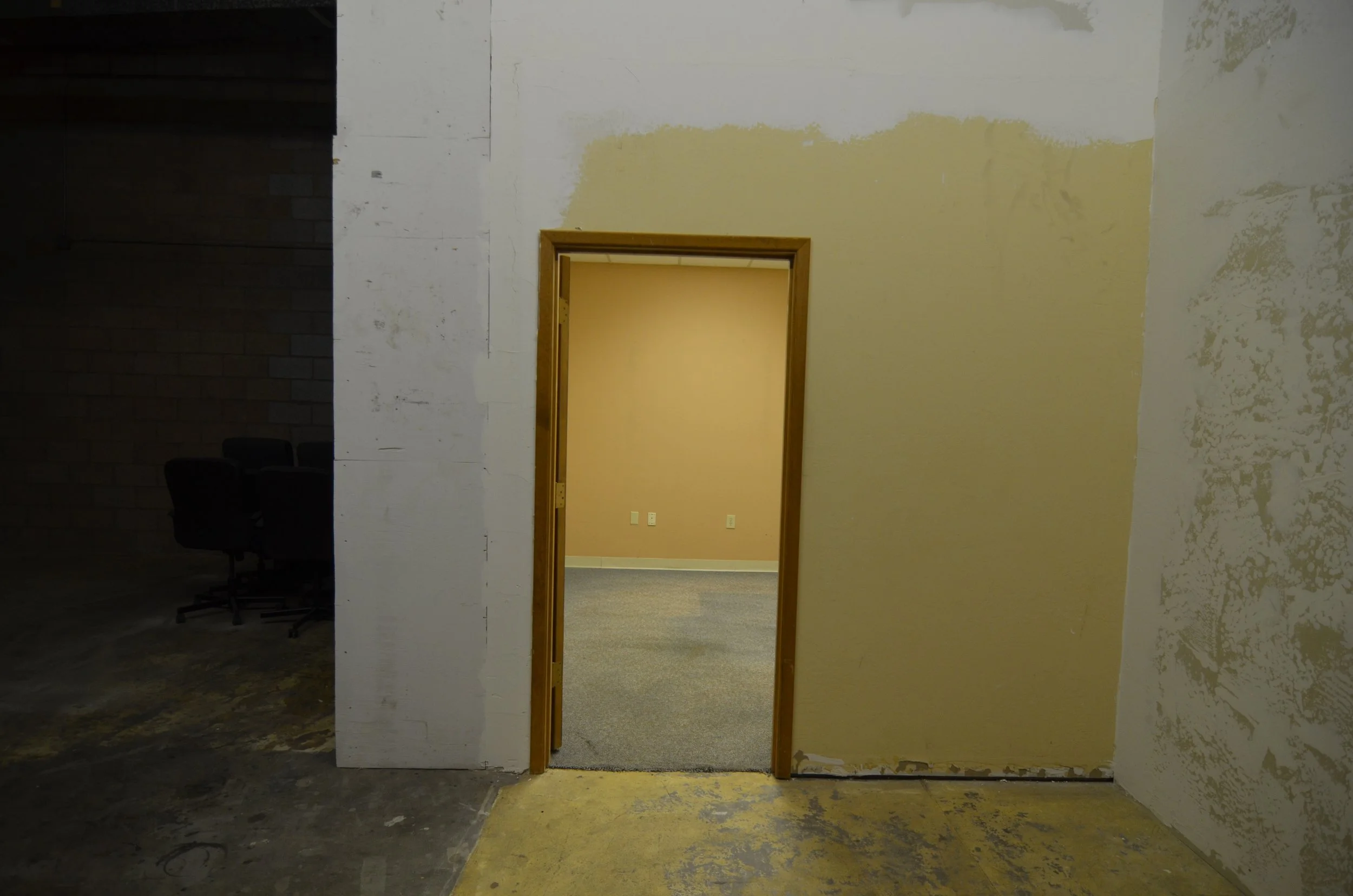 An unfinished interior room with a doorway leading to another room. The walls are partially painted and in need of repair, with an open space revealing a beige wall and gray carpeted floor inside.