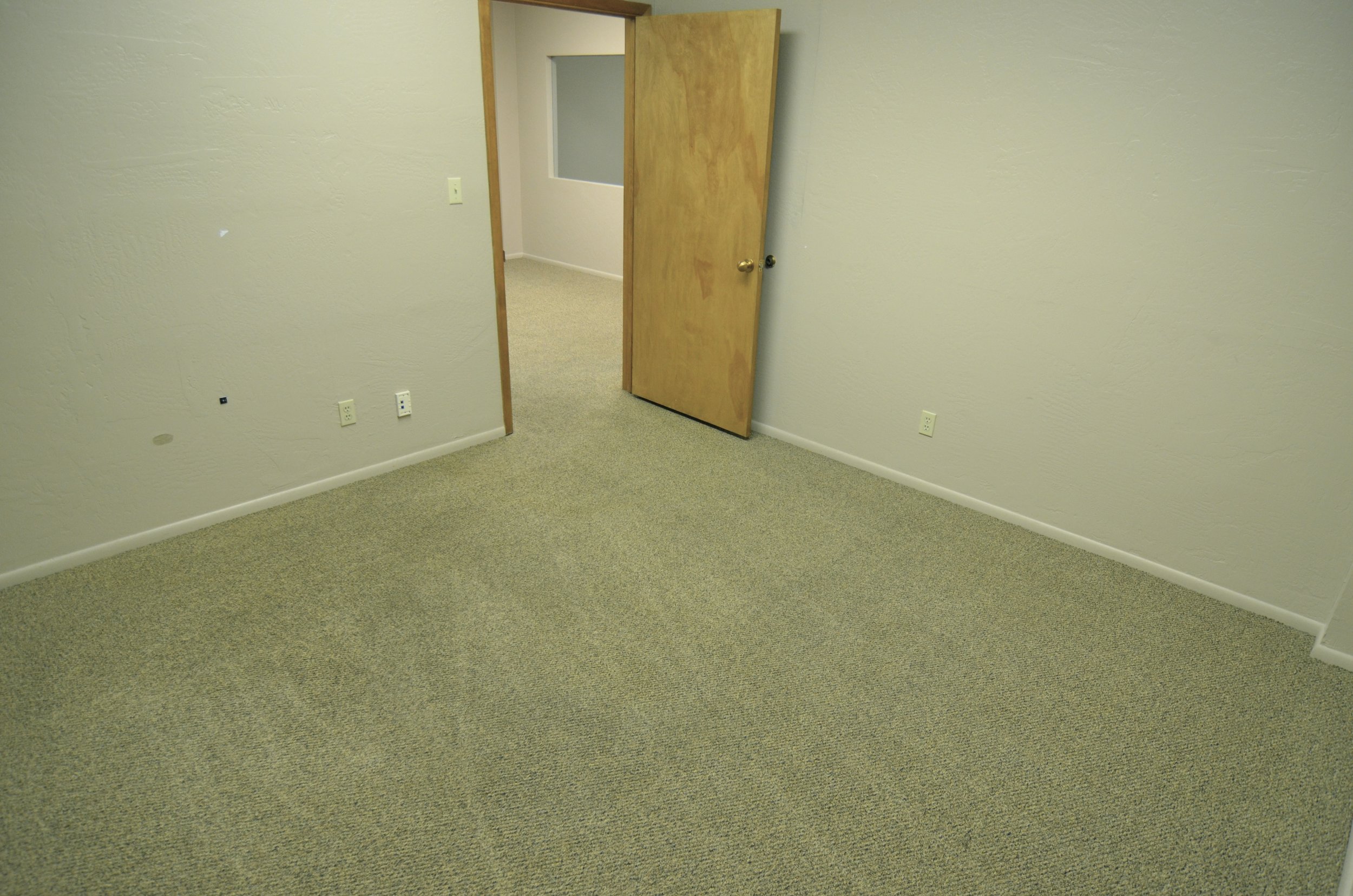 An empty room with beige carpet, plain off-white walls, and a wooden door that is partially open, revealing a small adjacent room or closet.