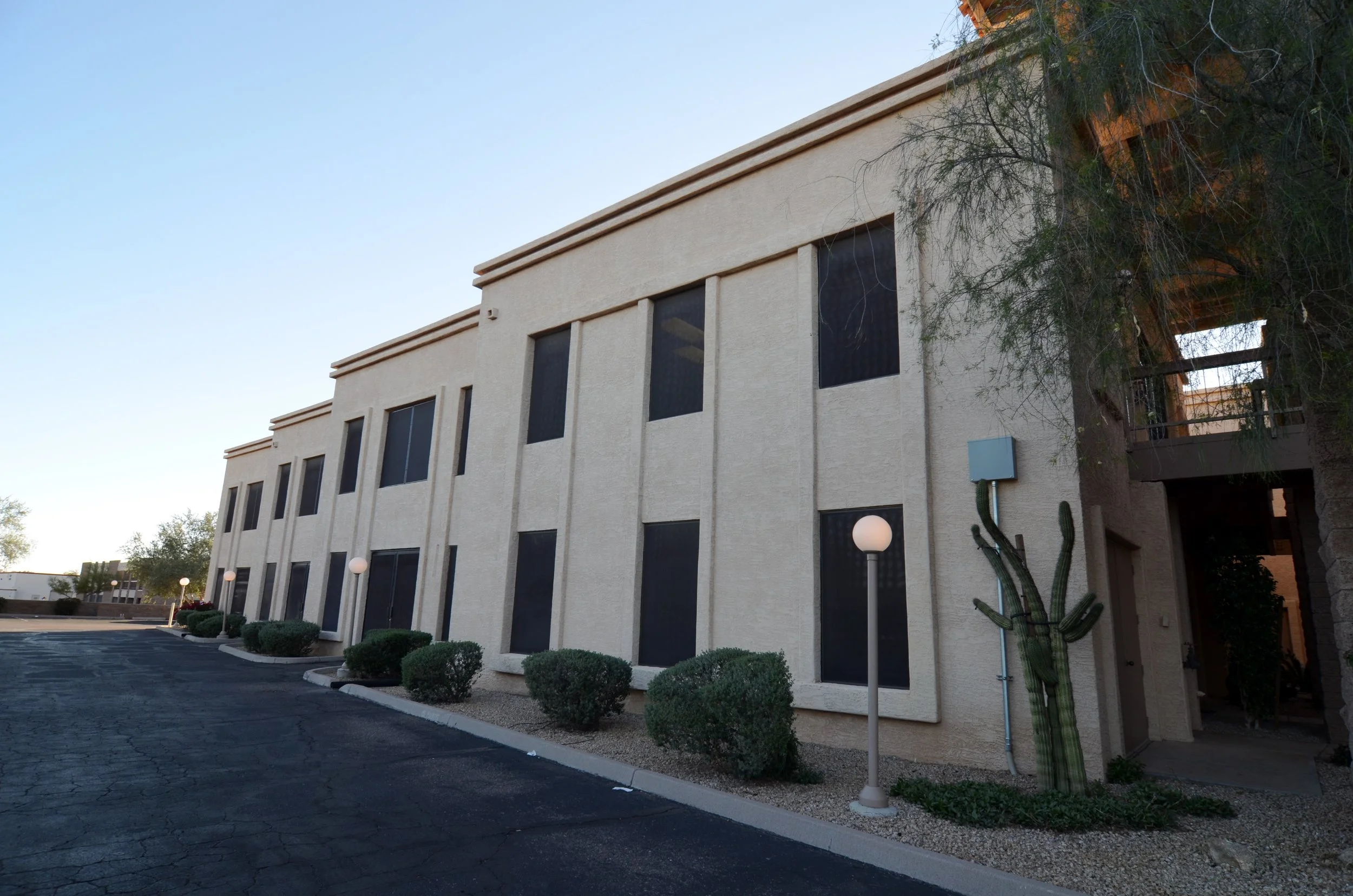 A beige stucco building with tall, narrow windows, a small landscaped area with bushes and a cactus, and vintage-style street lamps. A large tree partially obscures the building, with a clear sky overhead.