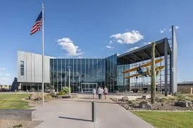 A modern building with large glass windows, an American flag flying on a flagpole, and a construction crane near the entrance, with a clear blue sky overhead.