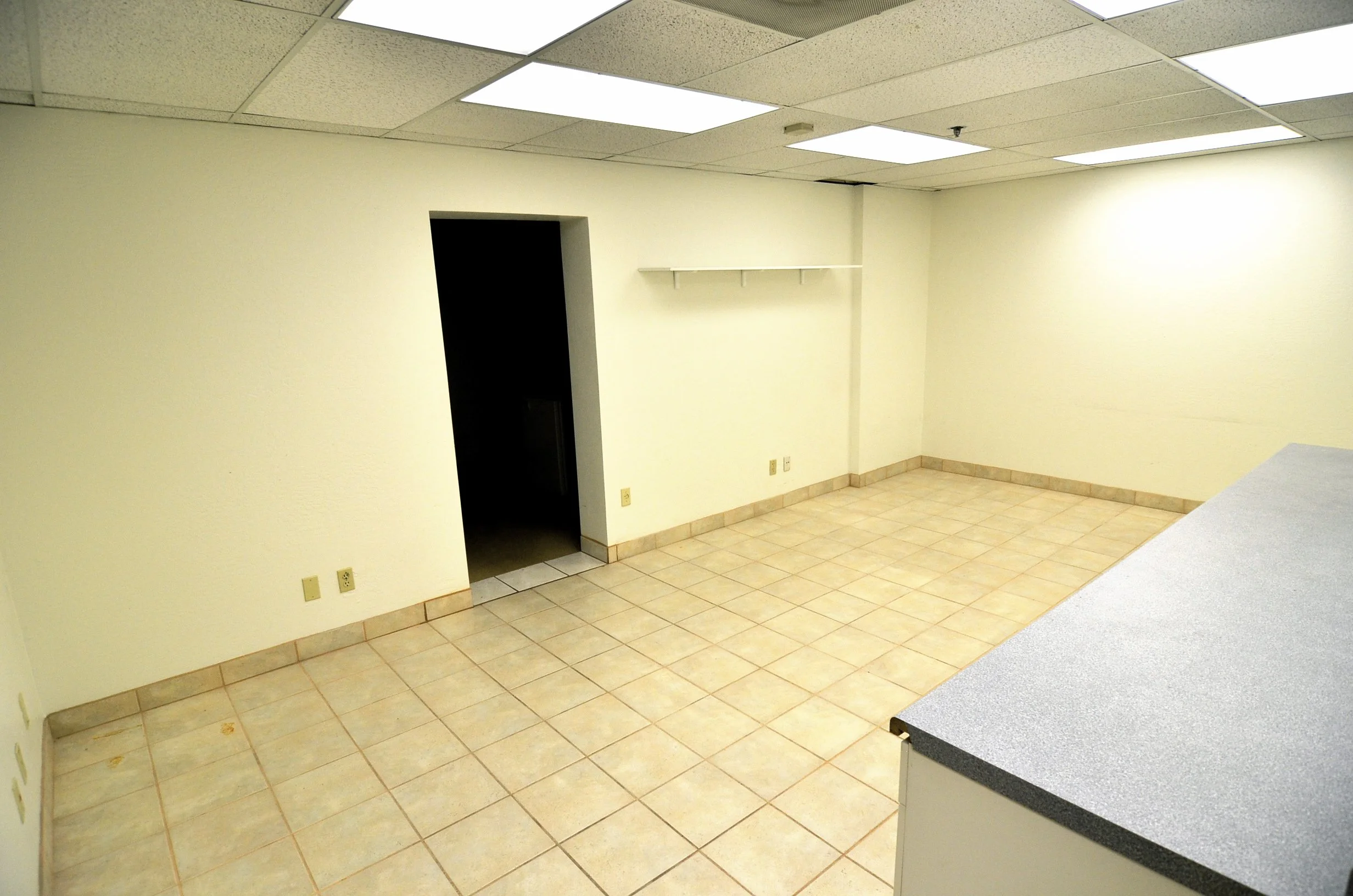 An empty room with yellow walls, beige tiled floor, and a white ceiling with fluorescent lights. There is a small black door and a white shelf mounted on the wall. Part of a black countertop with a gray granite surface is visible in the foreground.