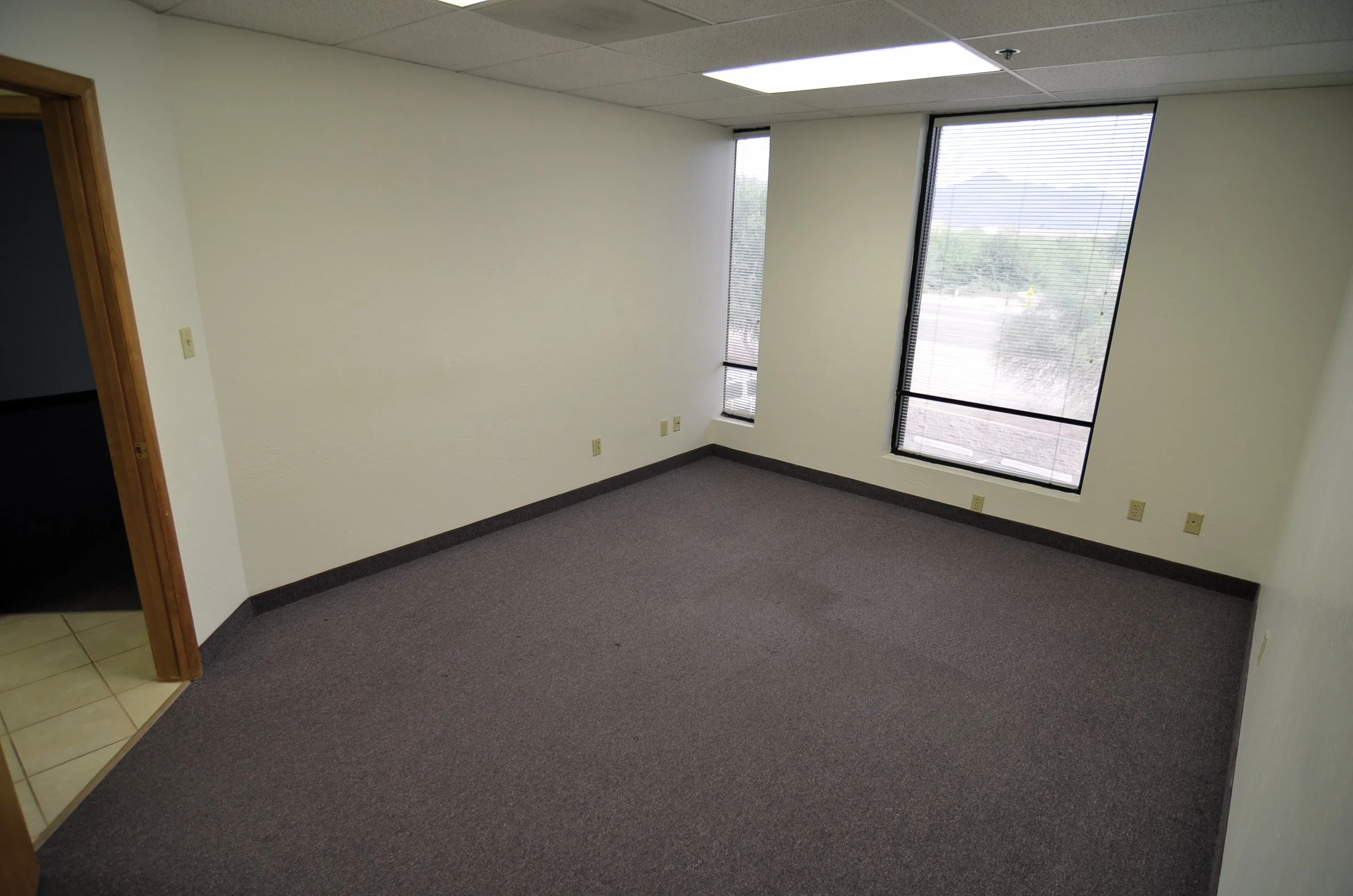 Empty office room with gray carpet, white walls, and large windows with blinds, revealing a parking lot outside.