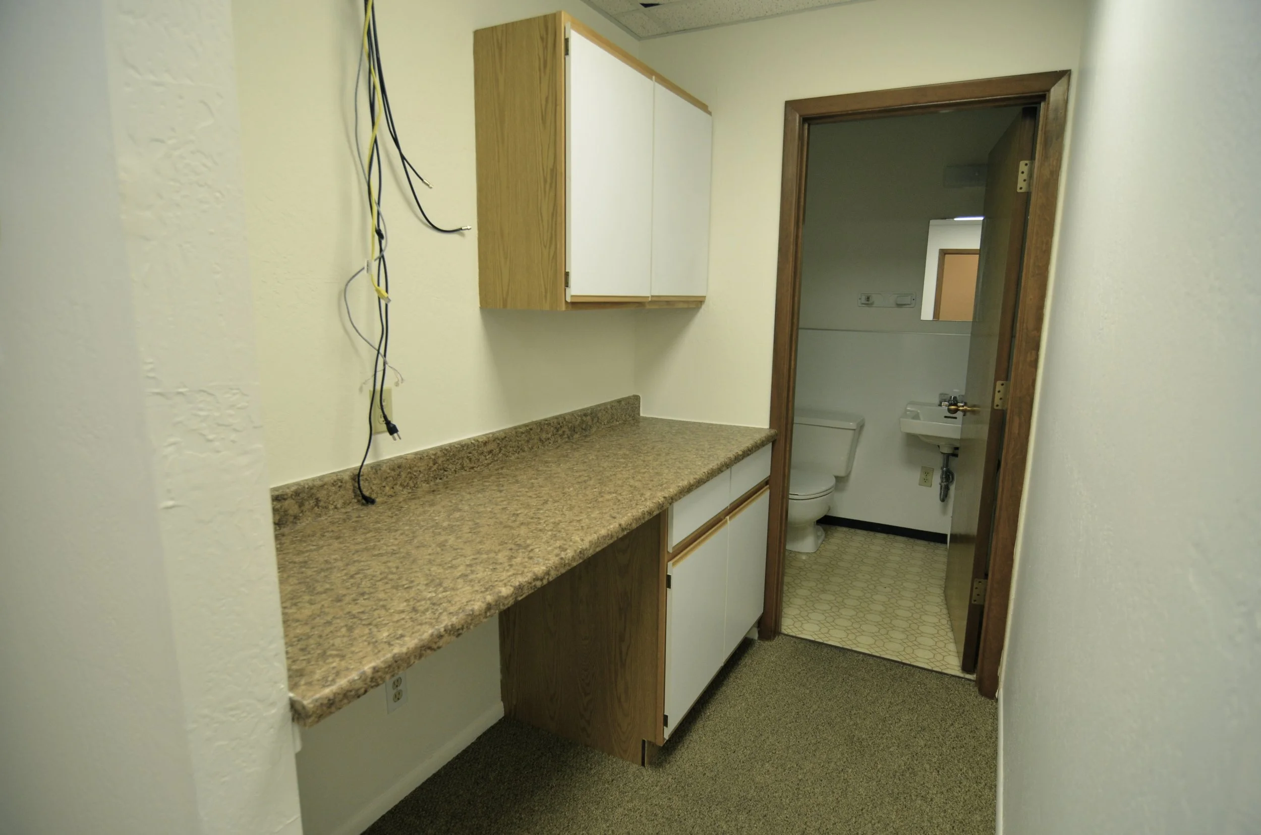 Empty office kitchenette with beige countertop, white cabinets with wooden trim, and a small bathroom with a visible toilet and sink through an open door.