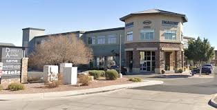 A two-story commercial building with a sign for Dental Care and other businesses, situated on a parking lot with trees and bushes nearby.