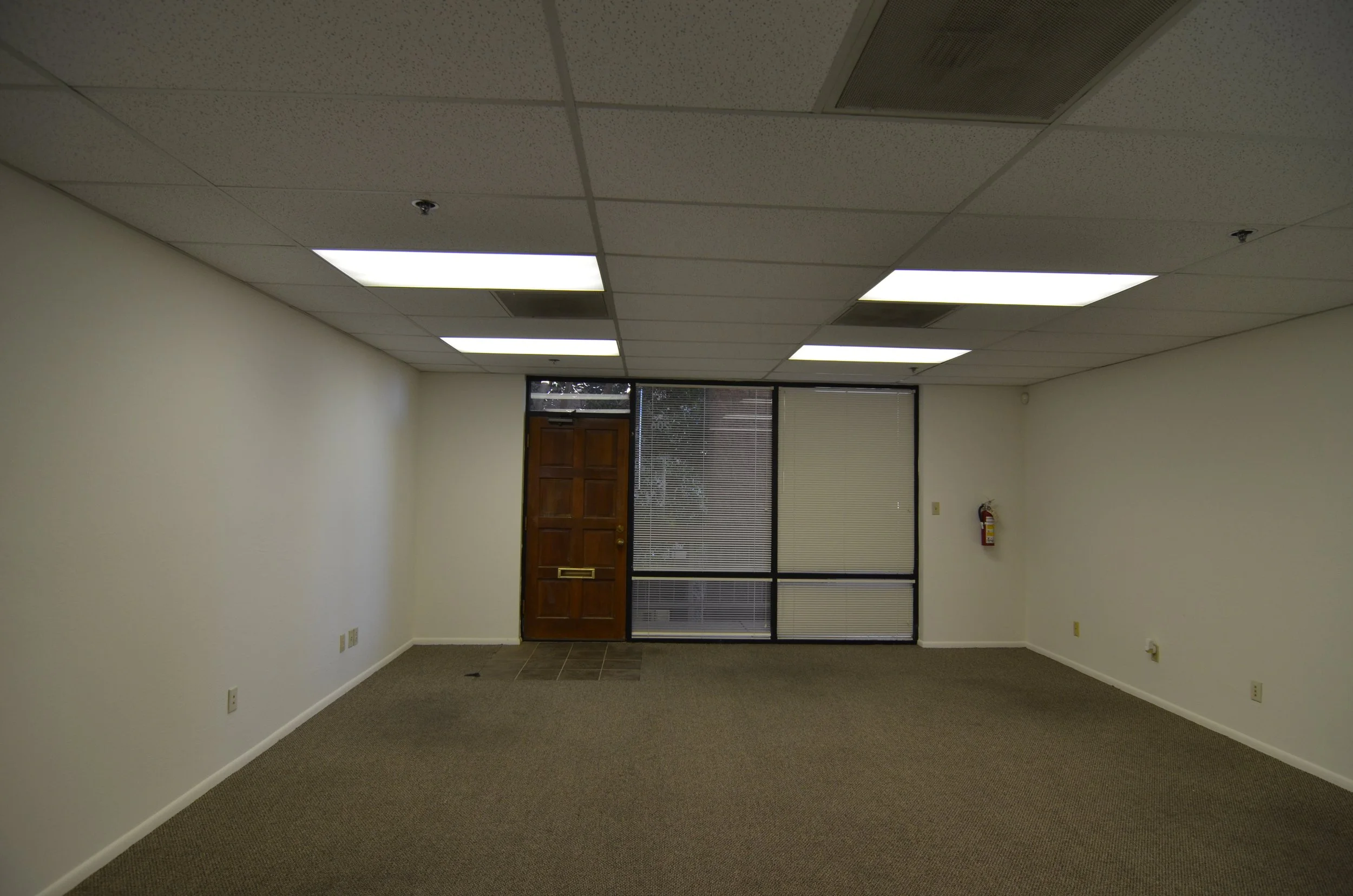 Empty office room with beige walls, gray carpet, ceiling tiles with fluorescent lights, a wooden door, and large windows with blinds.