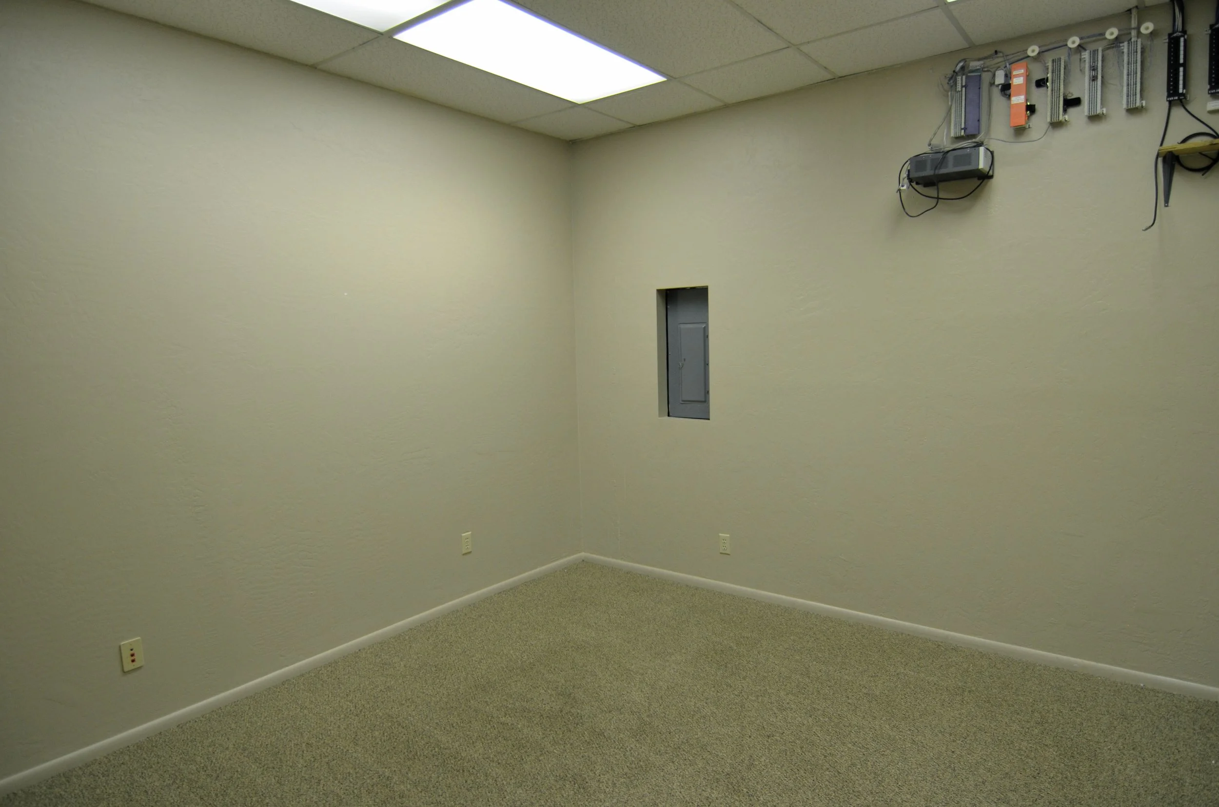 An empty office room with light beige walls, a beige carpet, a small electrical panel on the wall, ceiling tiles, and a large fluorescent ceiling light.