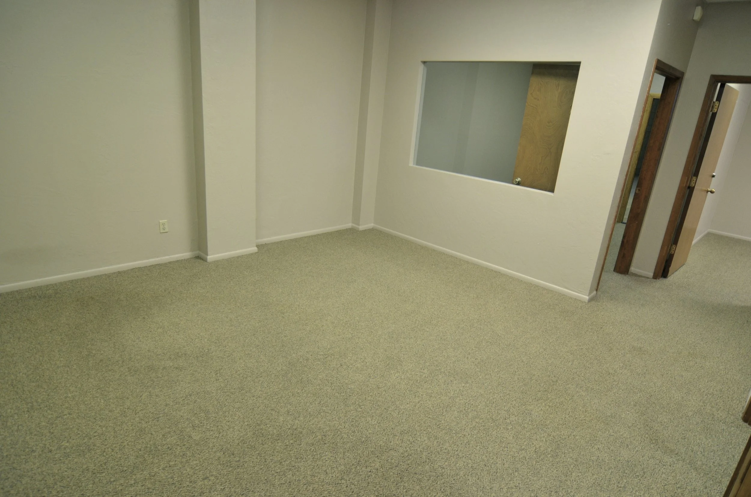Empty room with beige carpet, off-white walls, a small window, and a doorway with a wooden frame leading to a hallway.
