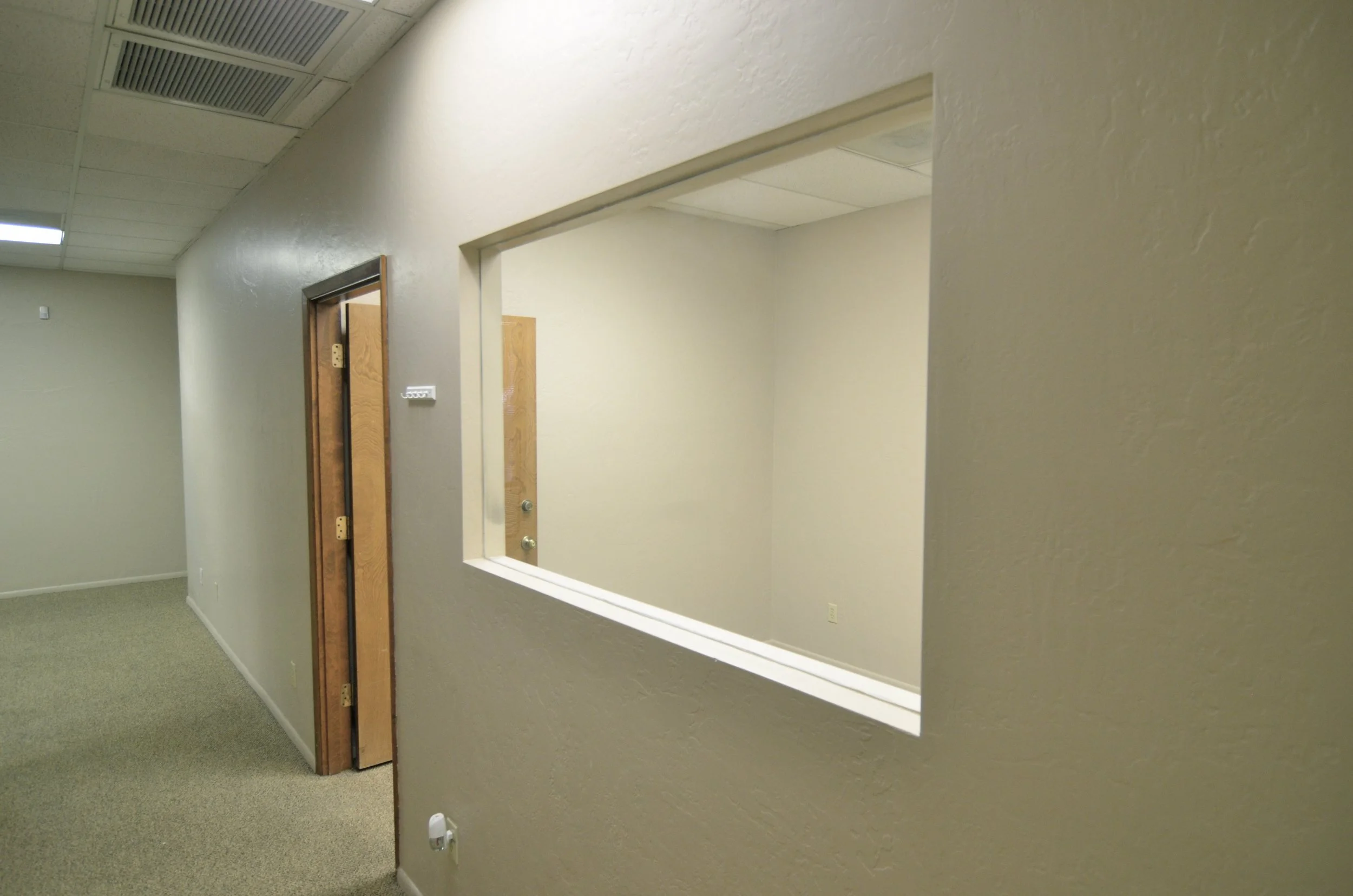 Interior view of an office or medical clinic, showing a hallway with beige walls, carpeted floor, ceiling with air vents, and a window in the wall overlooking another room with a wooden door.