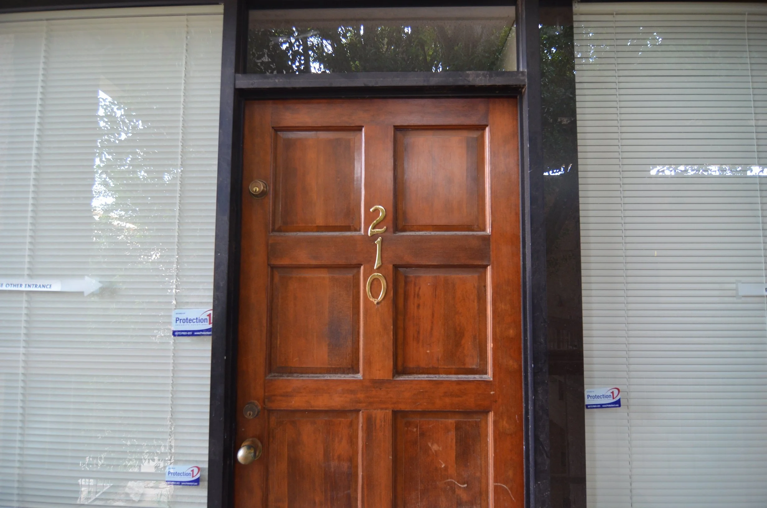 Wooden front door with the number 210 on it, set between large window panels, and blinds.
