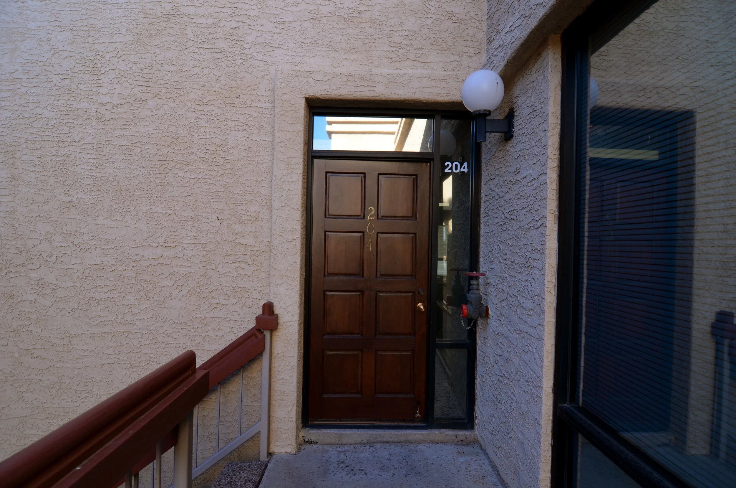 View of a residential front door with the number 204, brown wooden door, surrounding beige textured wall, light fixture above, and part of a window on the right.