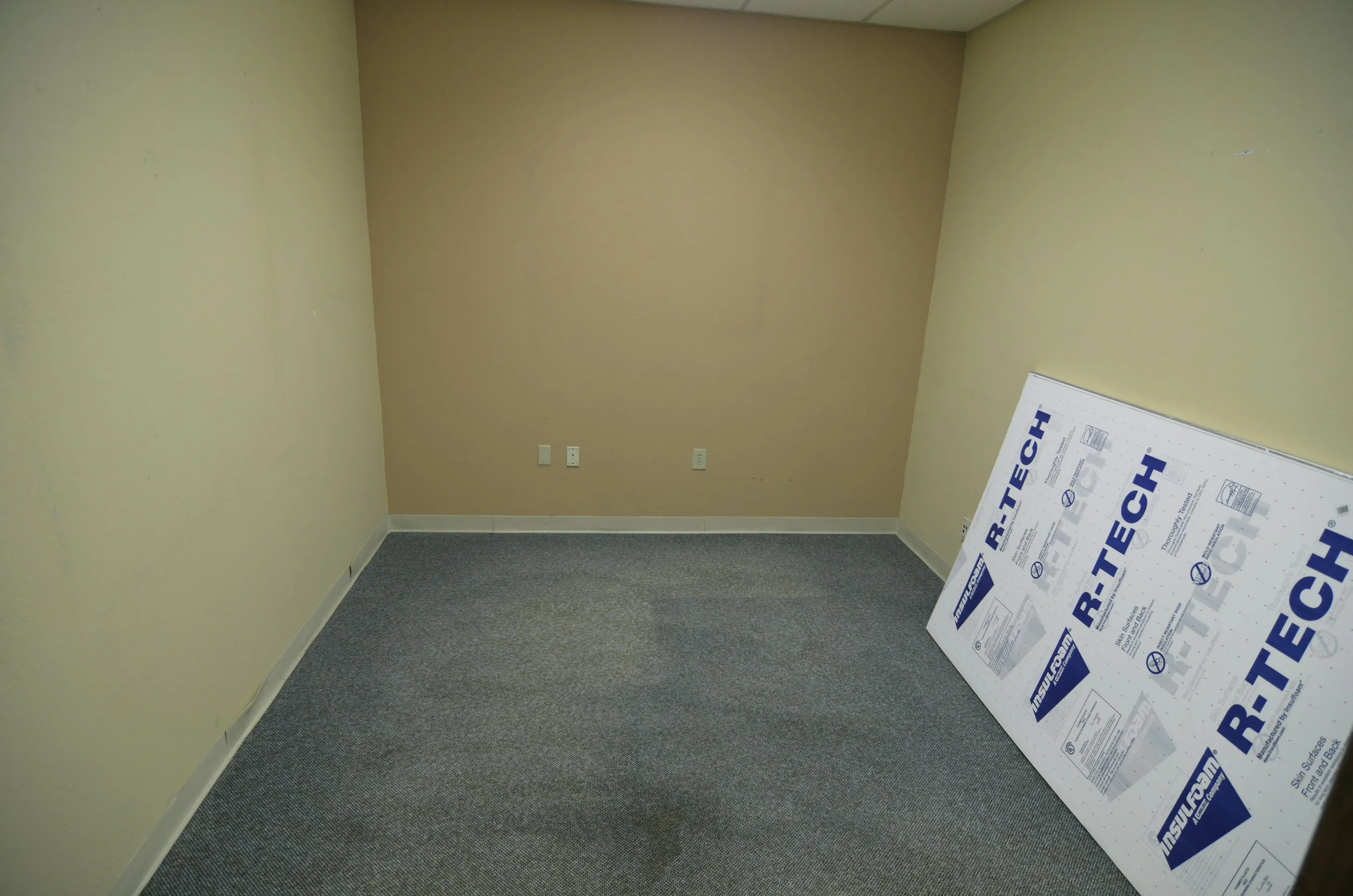 An empty beige-colored room with gray carpet, three wall outlets, and a piece of foam board leaning against the right wall.