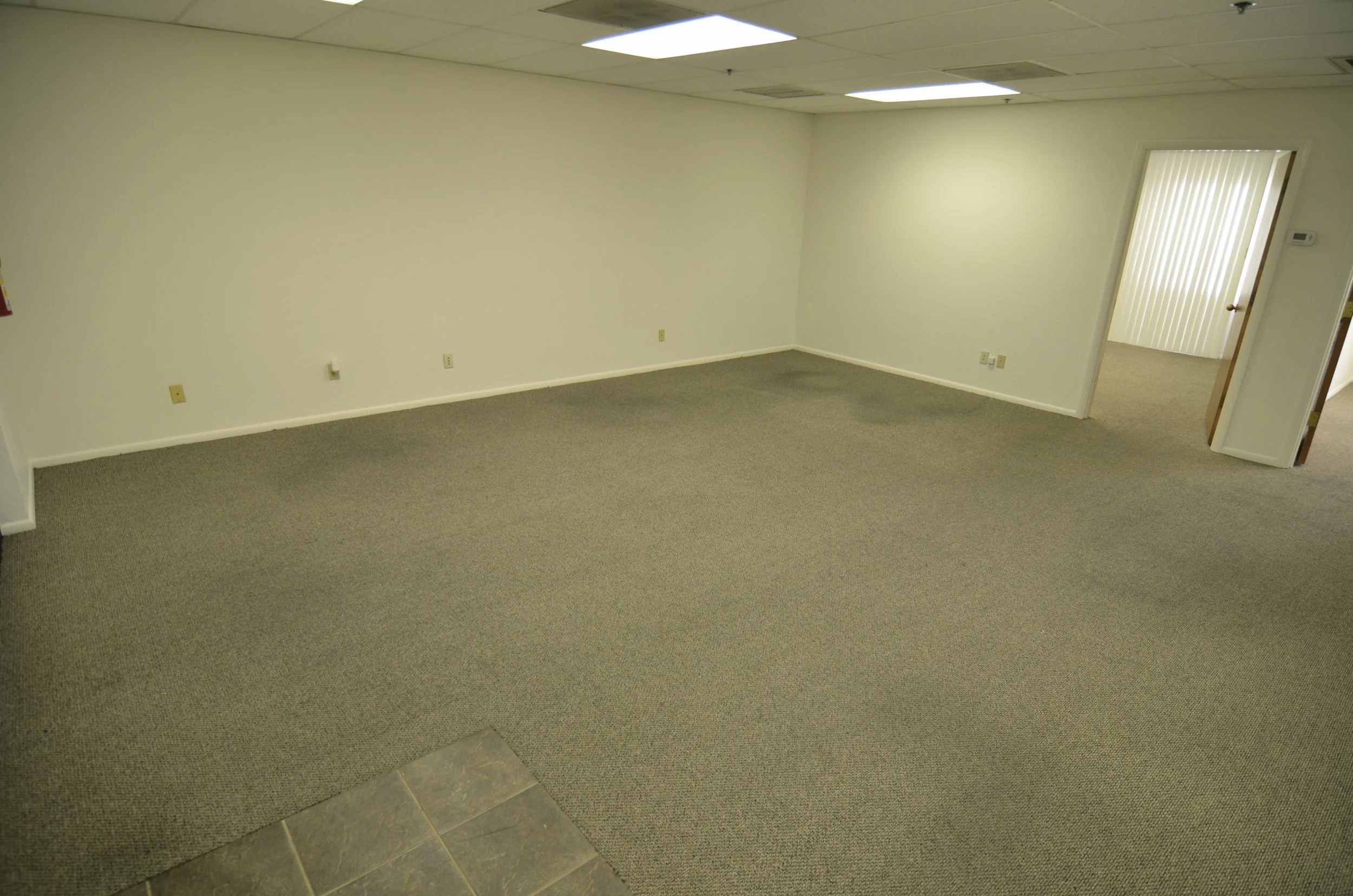 Interior of an empty office space with beige carpet, white walls, and a ceiling with fluorescent lights. There is a doorway leading to another room with vertical blinds and a thermostat on the wall.
