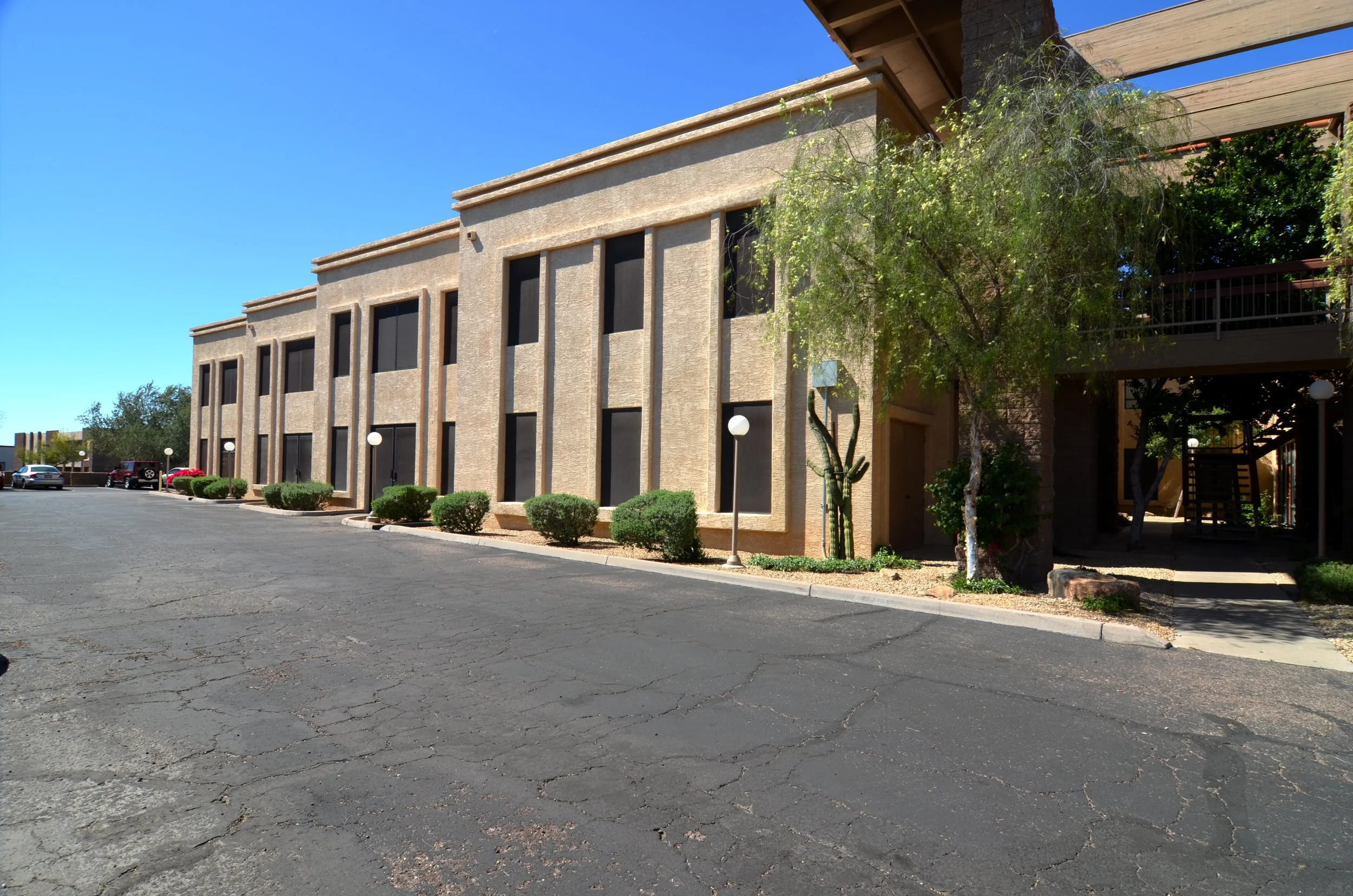 A two-story beige building with dark window screens, surrounded by desert landscaping, with parked cars and clear blue sky.