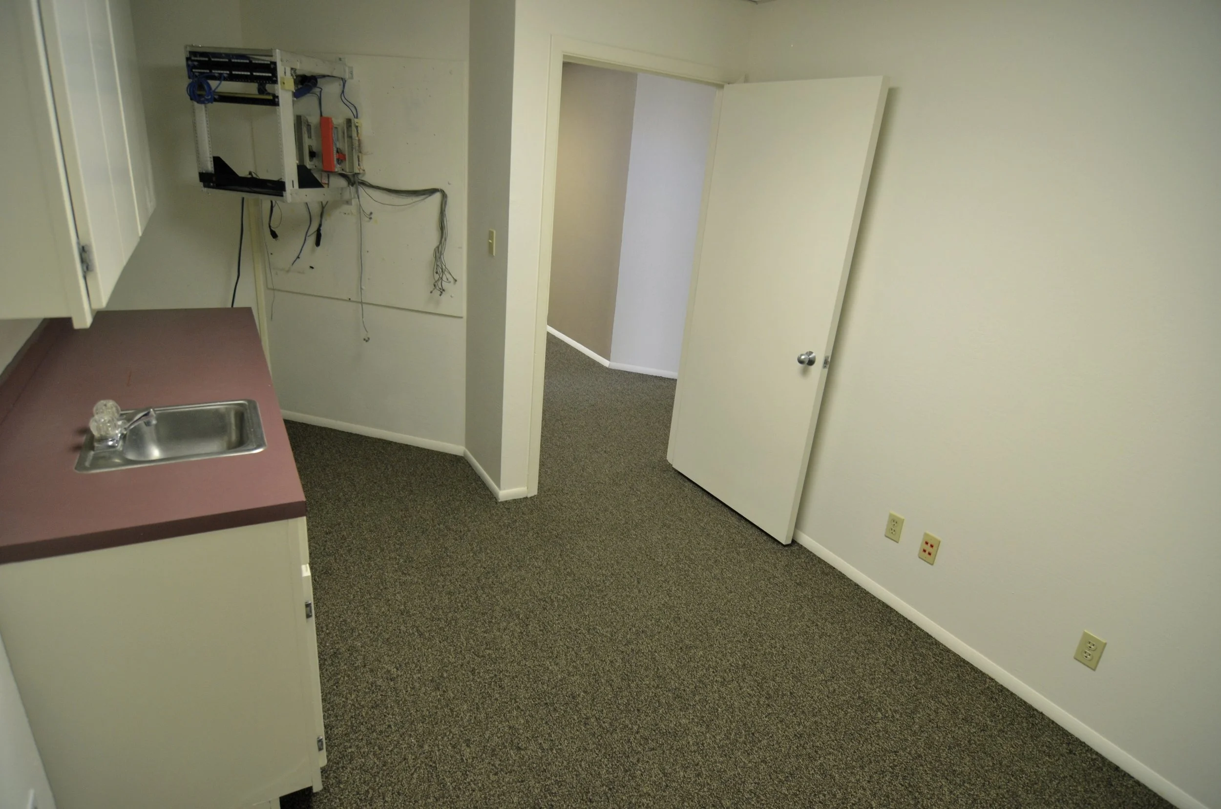 Empty room with a small kitchen area, including a stainless steel sink, pink countertop, and an open wall cabinet. There's an open door leading to another room or hallway, with some electrical wiring and an unfinished wall section exposed.