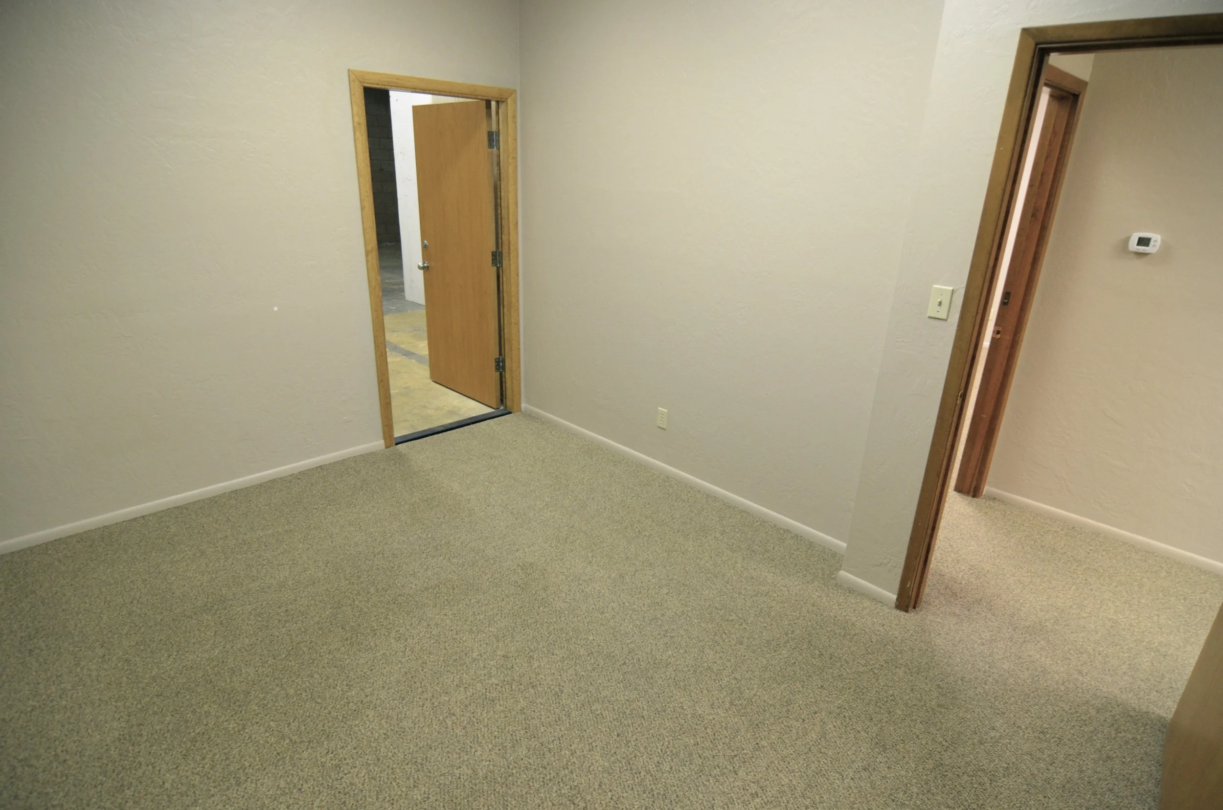 Interior of a room with beige walls, beige carpet, and two wooden doorways, with one door partially open revealing a hallway.