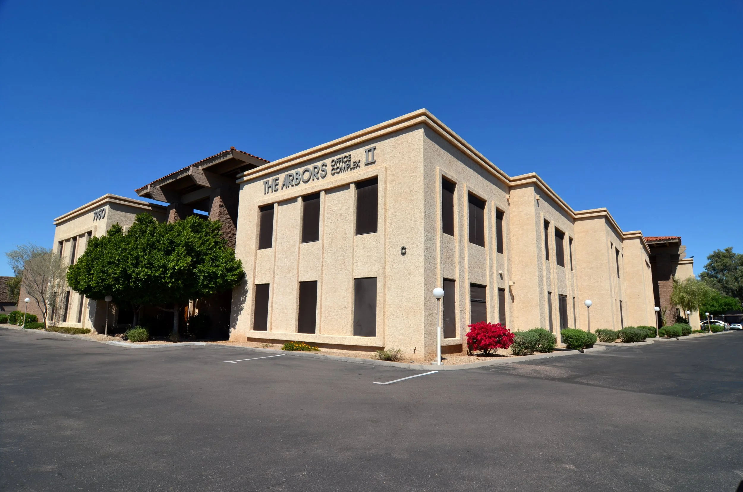 A beige commercial building with sign reading 'The Arbors Office Complex II', surrounded by small trees and bushes, under a clear blue sky.