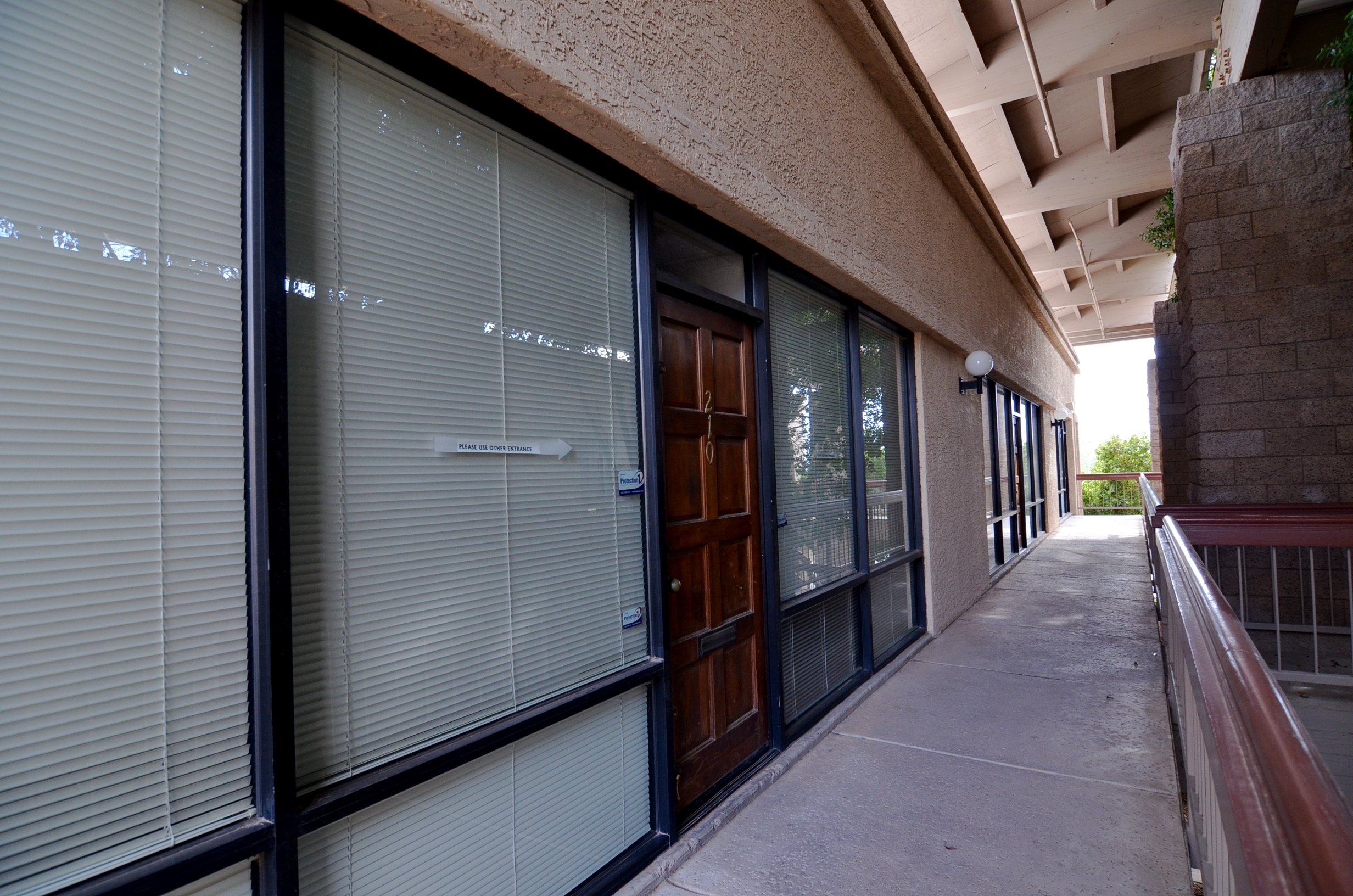 Office balcony outside entrance to Suite 210, with glass windows, beige stucco wall, wooden door with number 210, and outdoor lighting fixtures.