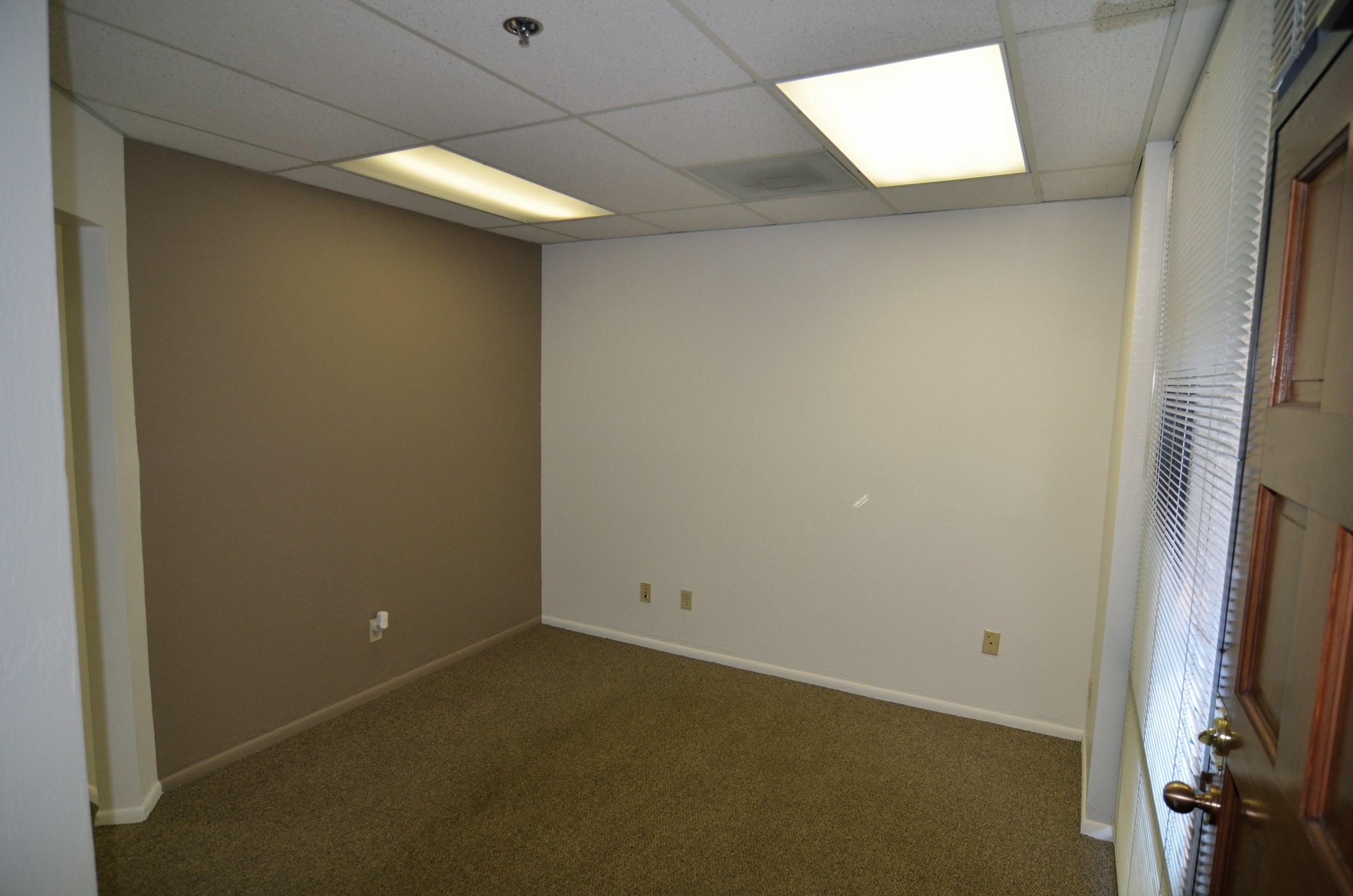 Empty room with beige and white walls, fluorescent ceiling lights, a window with closed blinds, flooring carpeted in brown, and corner electrical outlets.