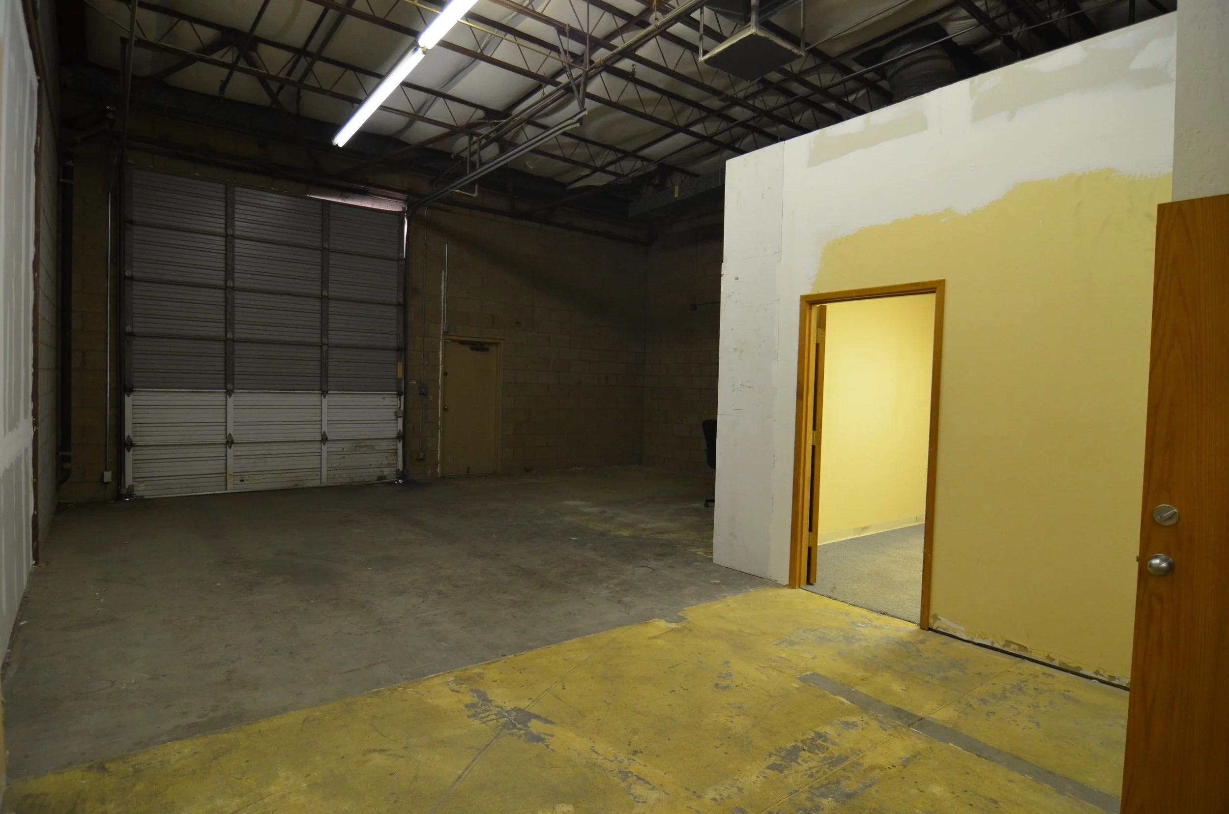 Interior of a warehouse or industrial space with a large silver roll-up door, concrete walls, and a partially finished room with yellow painted floors and an open door.