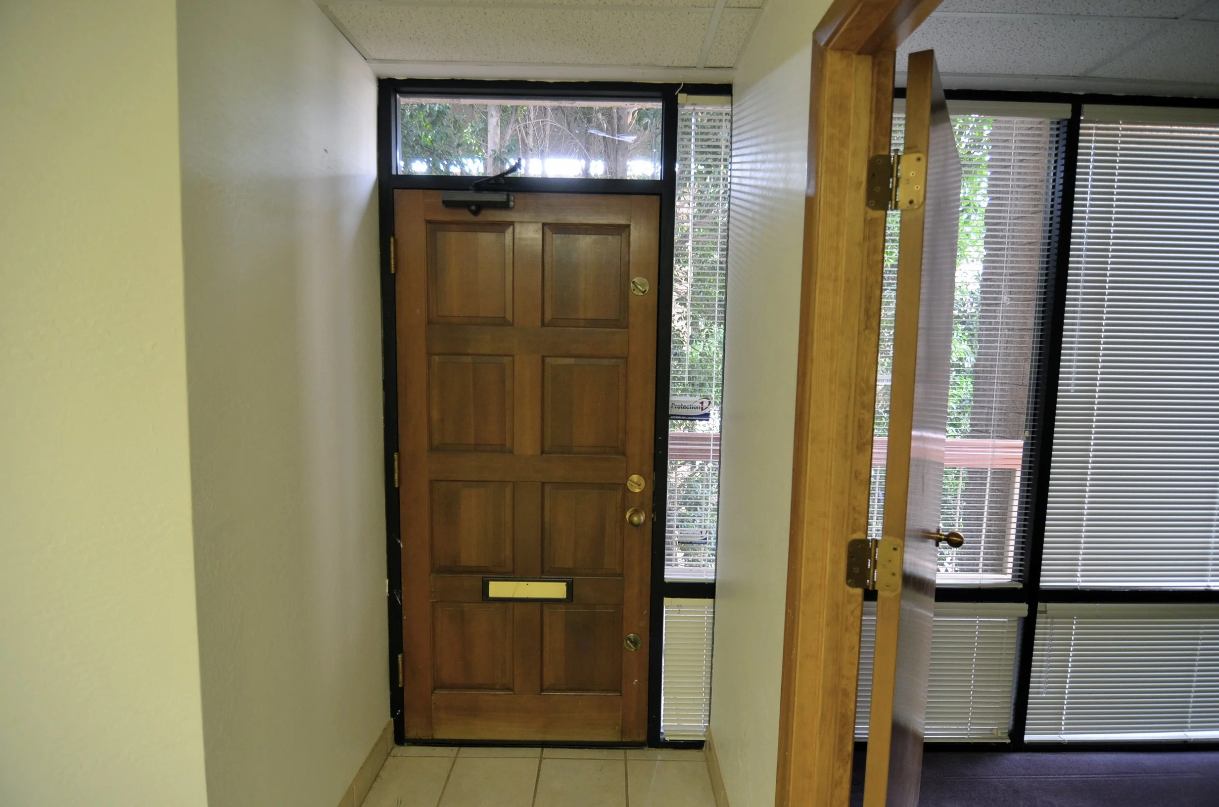 A wooden front door with a mail slot, surrounded by windows with blinds on either side. There is a hallway with light-colored tiled flooring and a wooden counter or bench in front of the window on the right.