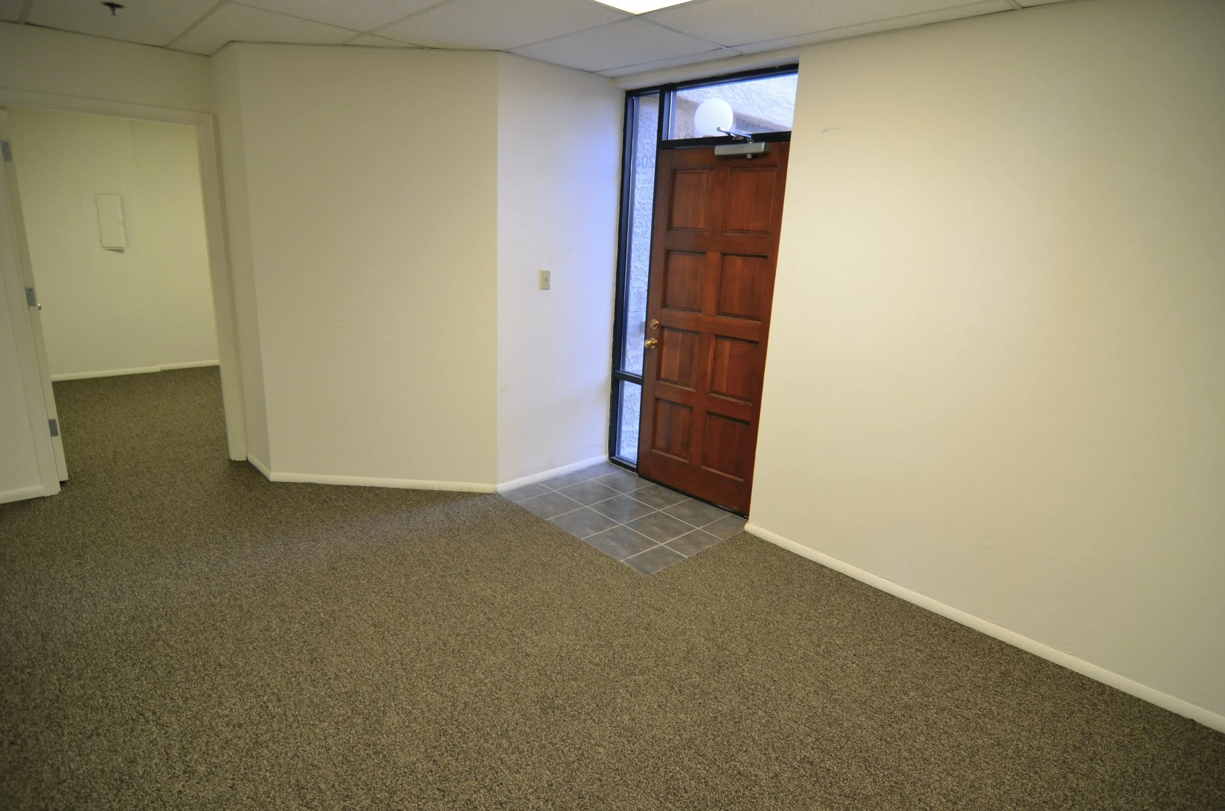 An interior view of a room showing a wooden front door with a glass panel, a doorbell, a small window above the door, and a plain light-colored wall. Part of the room has a carpeted floor and part has tiled flooring near the door.