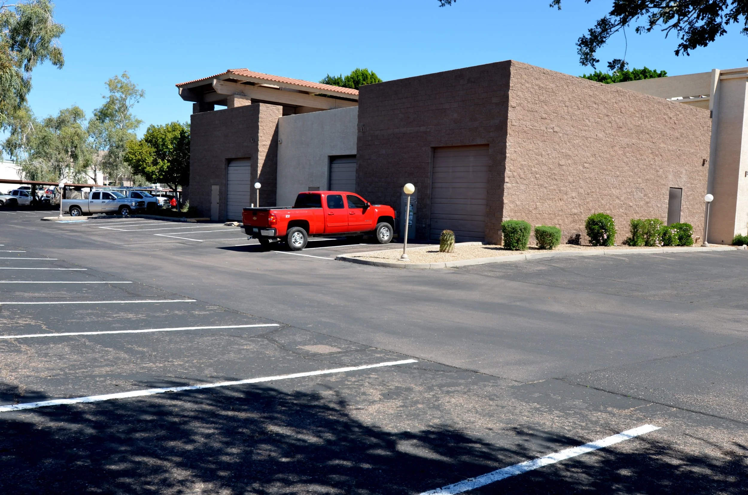 Empty parking lot with a red pickup truck, surrounded by a building with brown walls, trees, and blue sky.