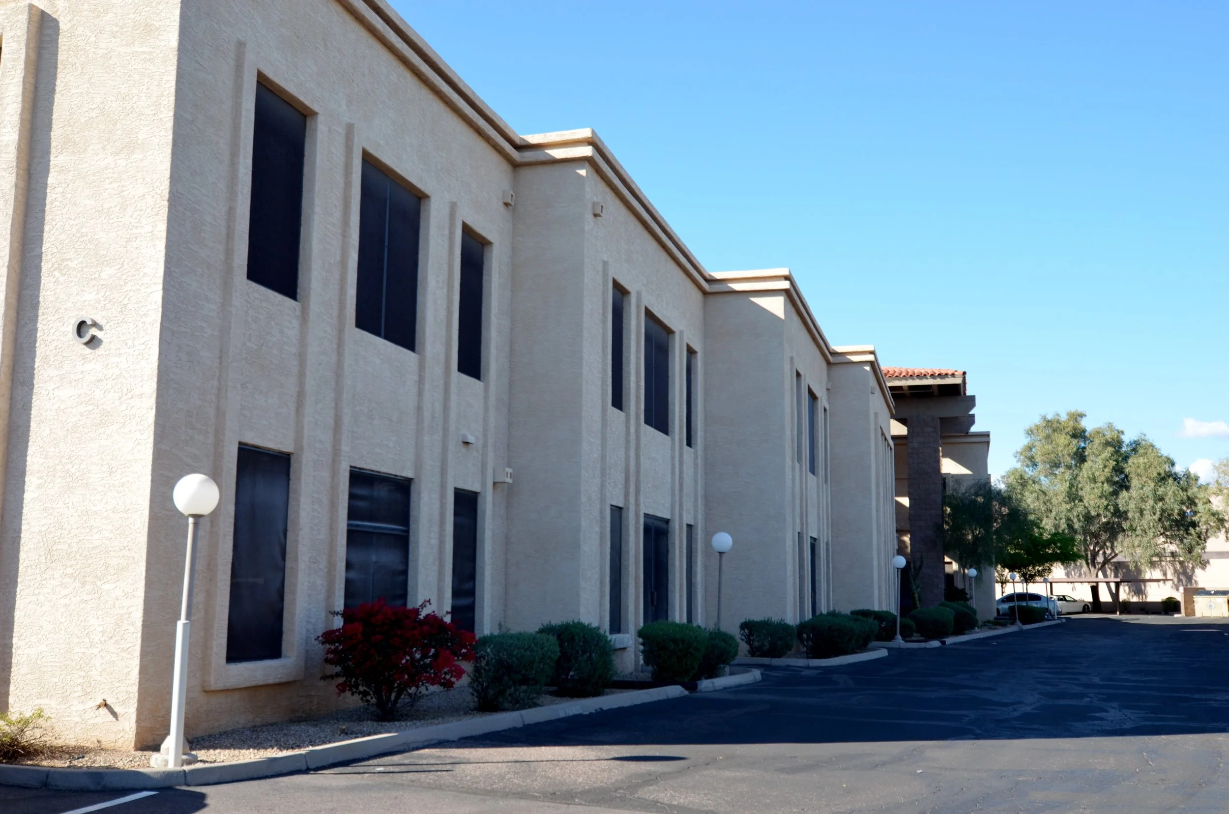 Exterior view of a beige multi-story building with a parking lot in the foreground, under a clear blue sky, with trees visible in the background.