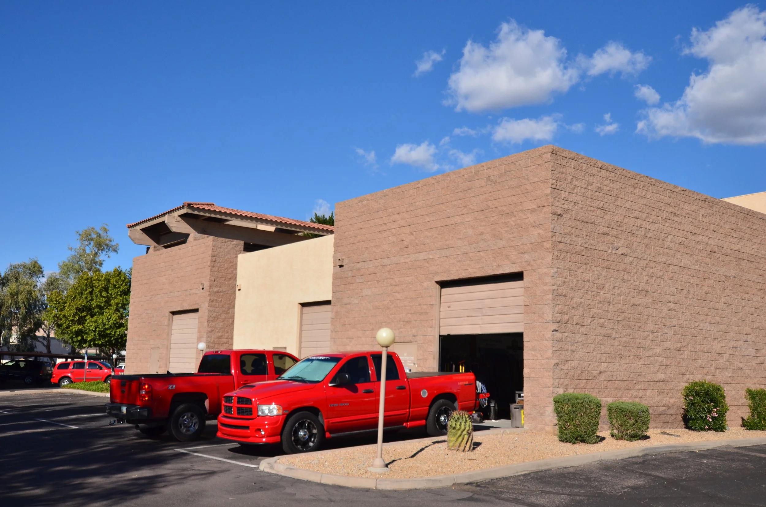 A modern beige brick building with several closed garage doors, parked red trucks, a parking lot, and desert landscaping in front of a blue sky with some clouds.