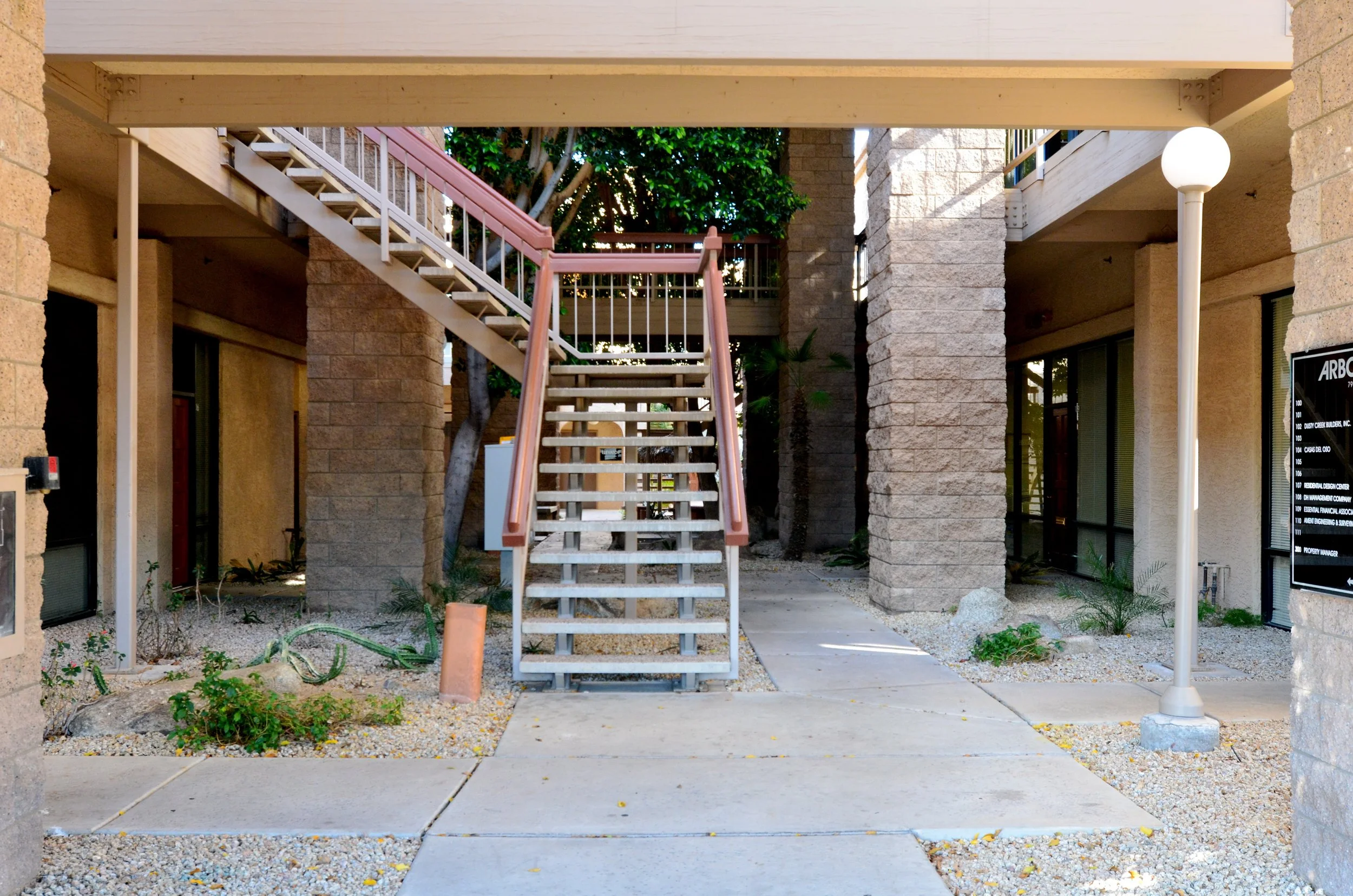 A view of an outdoor staircase leading to an upper balcony in a beige brick apartment complex, with a landscaped area below and a white lamppost on the right.