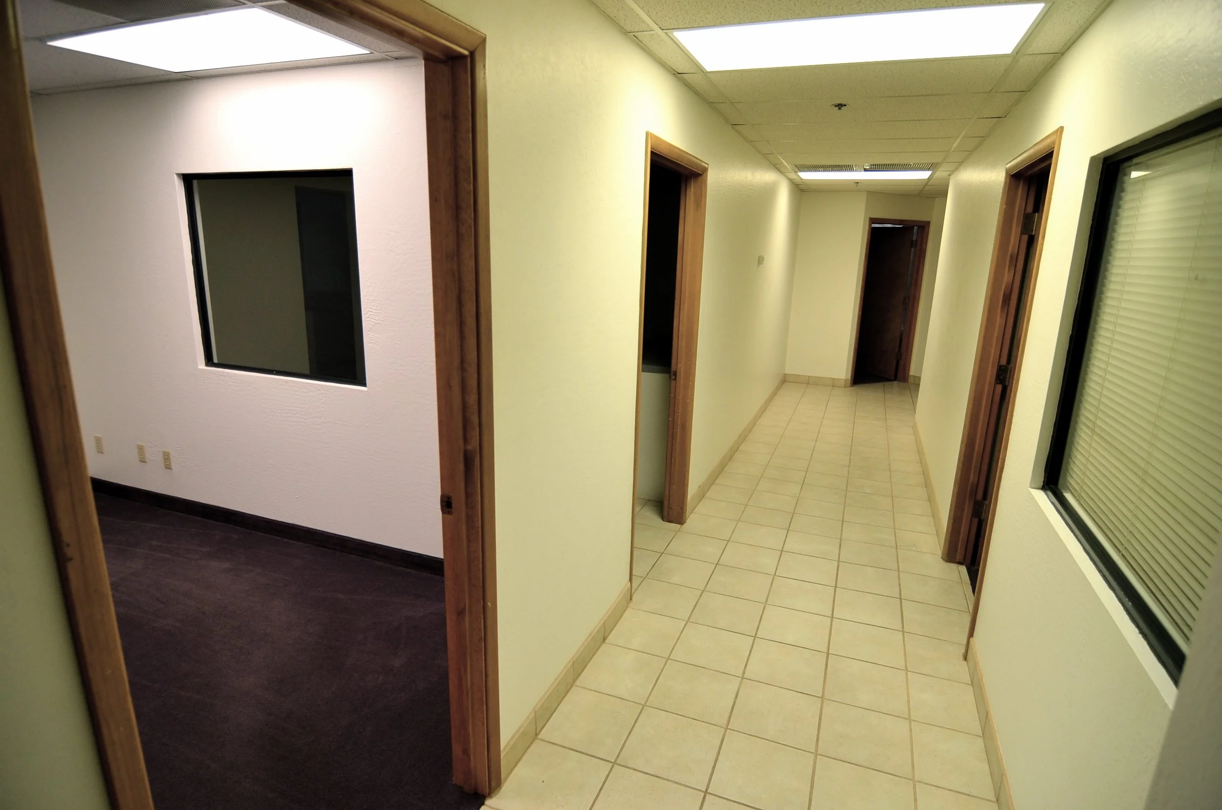 An office hallway with multiple doorways, tiled floors, and fluorescent ceiling lights. One doorway on the left leads to a room with dark flooring, while the other has light-colored walls with Venetian blinds on the window.