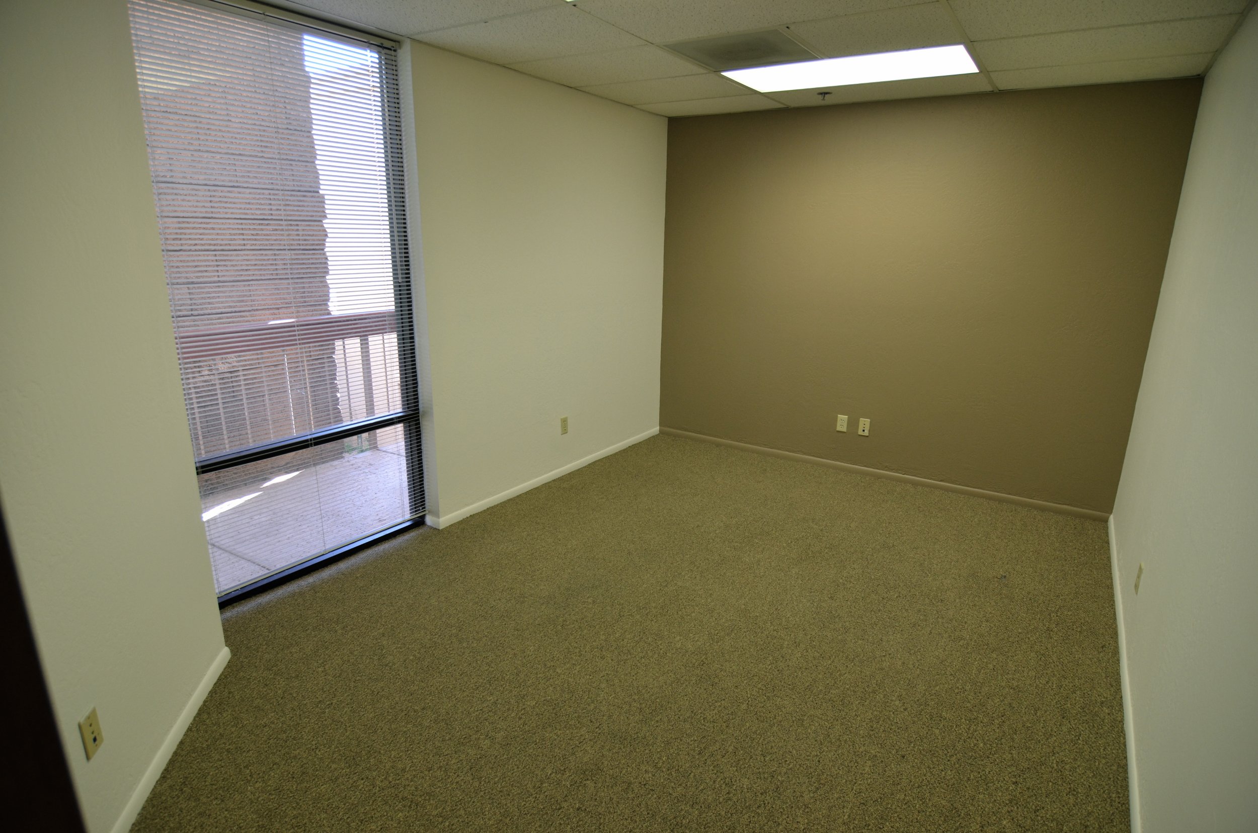 Empty office space with beige carpet, cream-colored and tan walls, a large window with closed blinds, and ceiling lights.