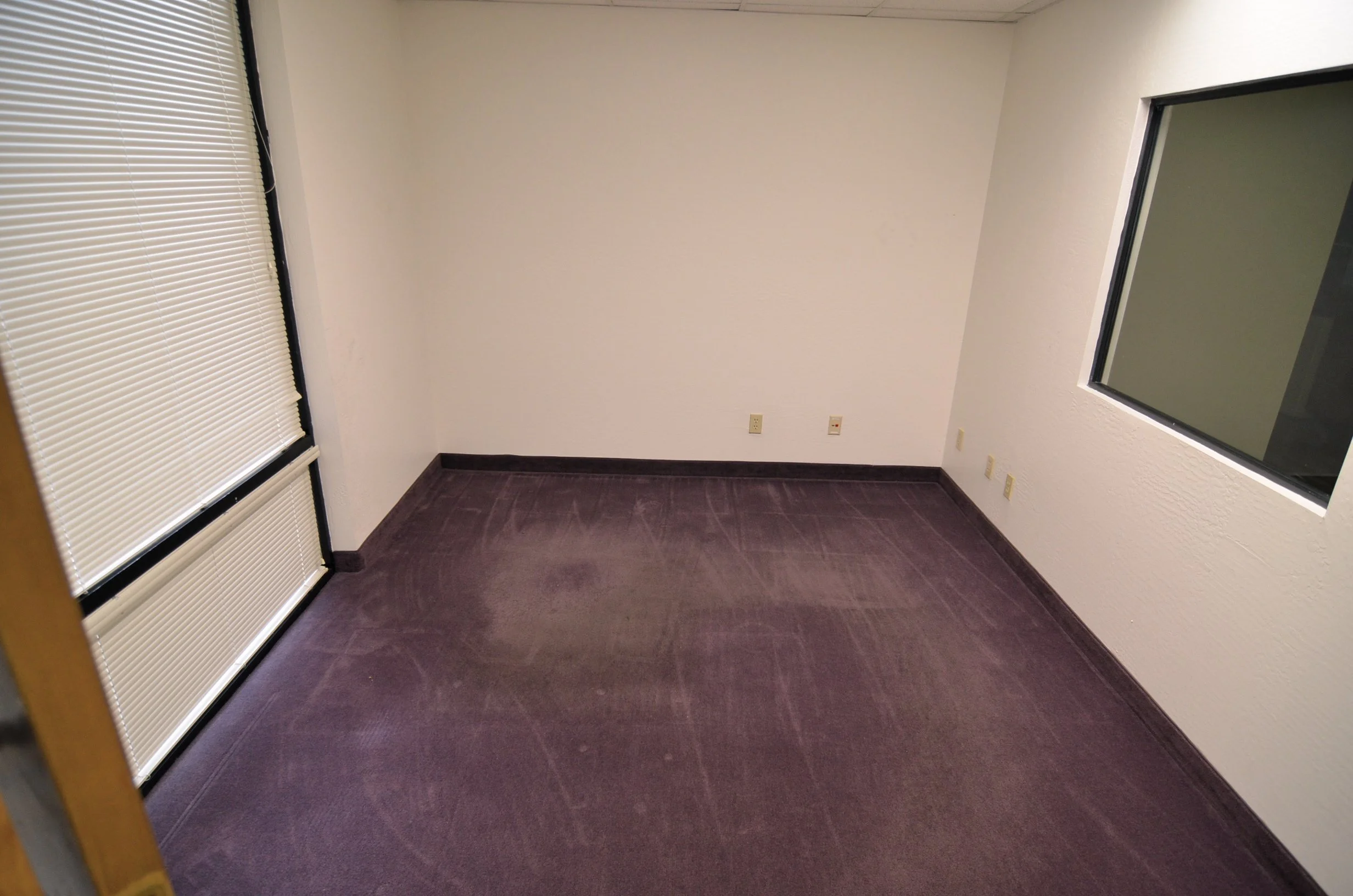 A small, empty office room with beige walls, purple carpet, a large window with closed blinds, and a glass window on the right wall.