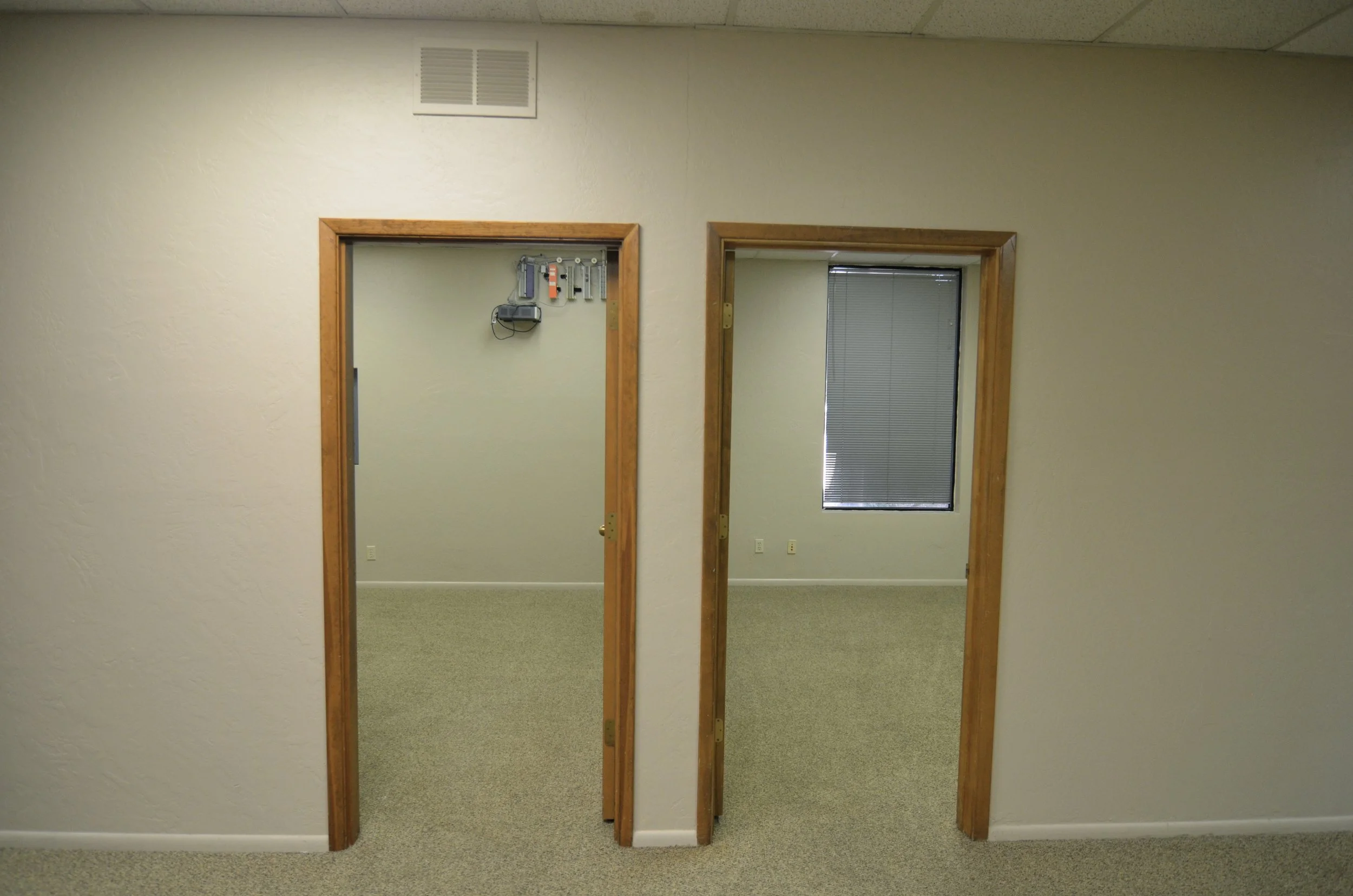 Interior view of a room with two doorways framed in wood. The left doorway shows a part of a room with tools on the wall and some electrical equipment, the right doorway leads to a room with a window and horizontal blinds. The wall is beige with a te