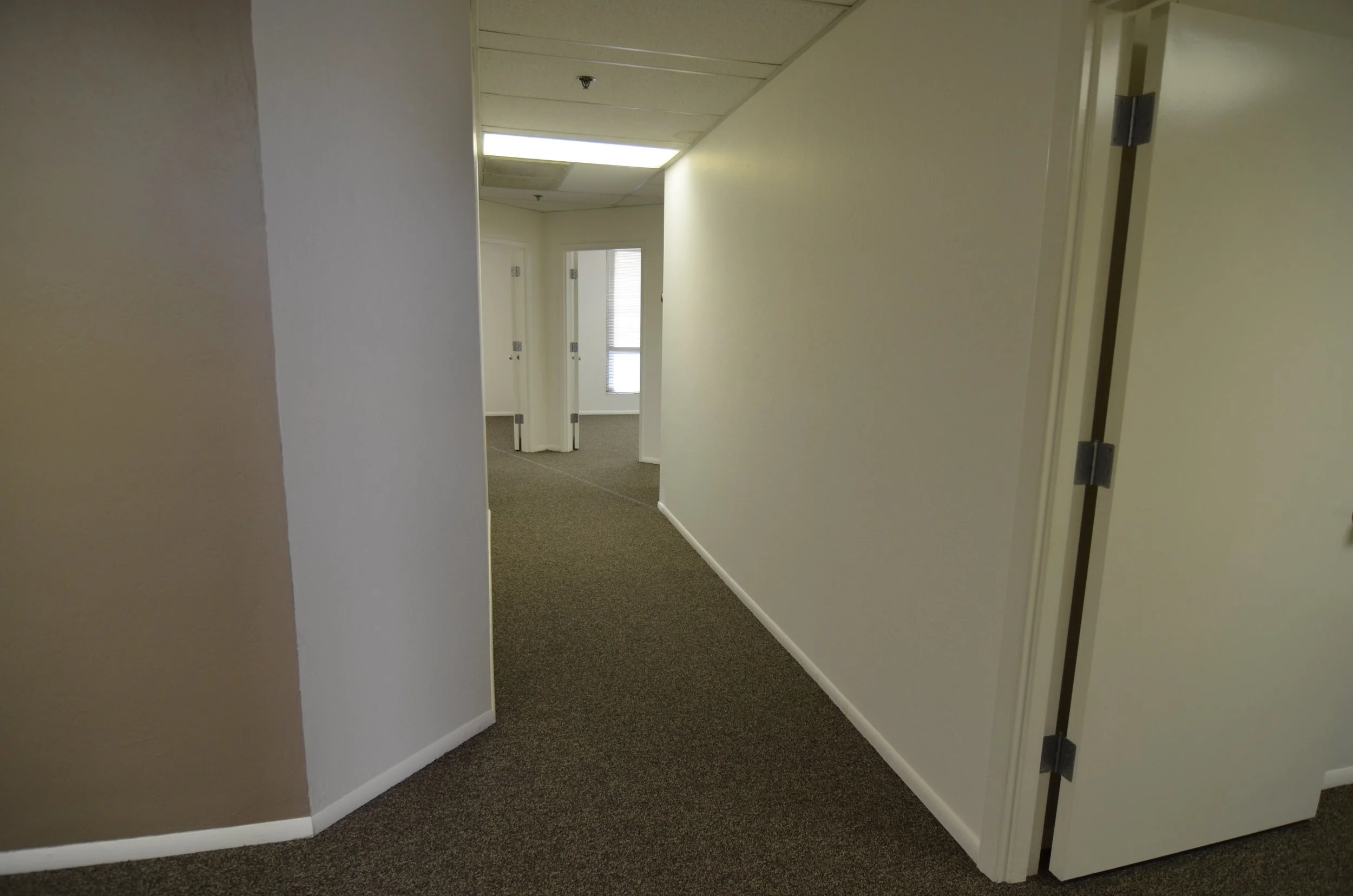 Office hallway with beige walls, dark carpet, and open doorways leading to other rooms. Fluorescent lighting overhead.
