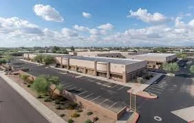 An empty parking lot in front of a large building with a beige facade, set under a partly cloudy sky.