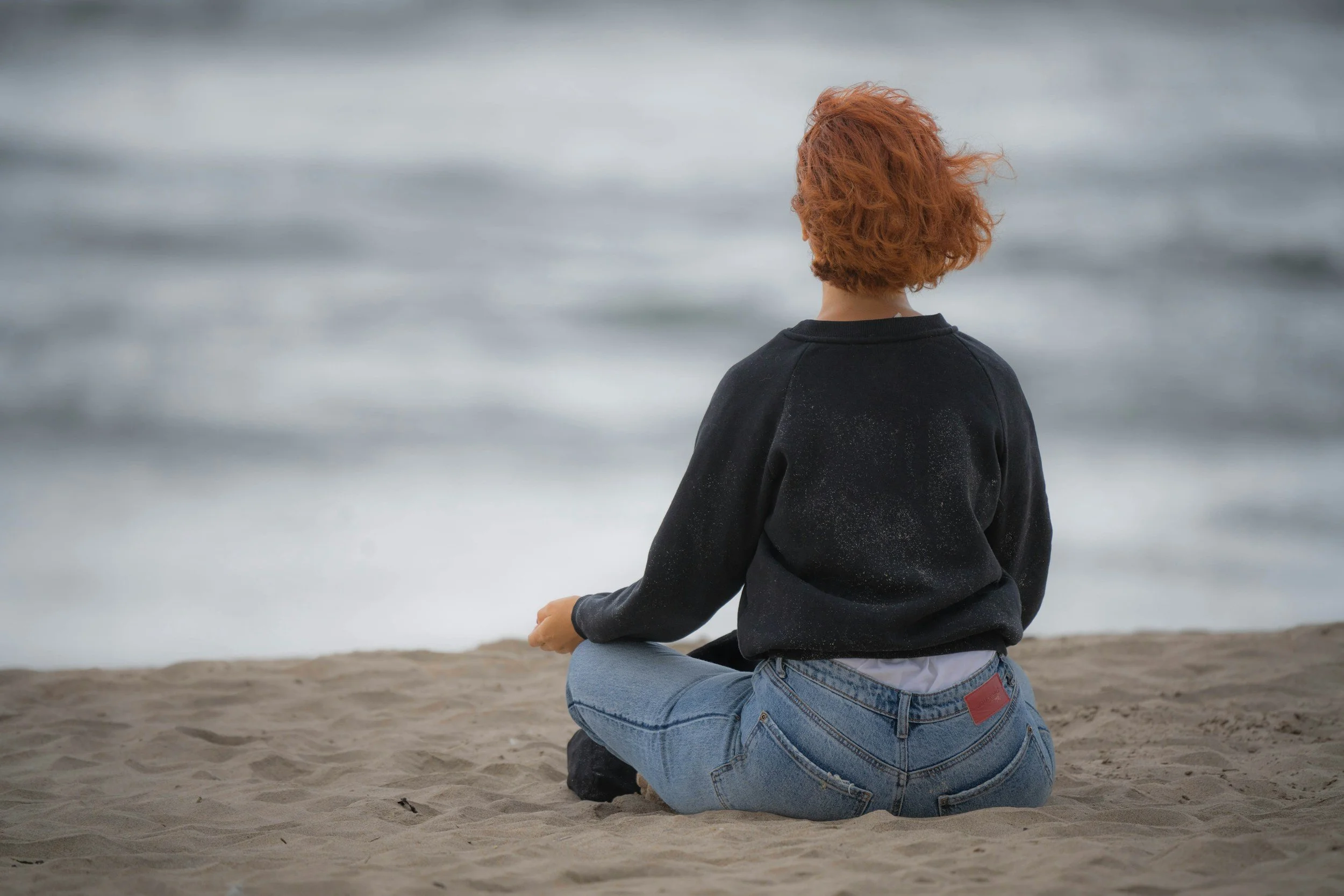 Junge Frau mit roten Haaren sitzt am Strand und blickt auf das Meer.