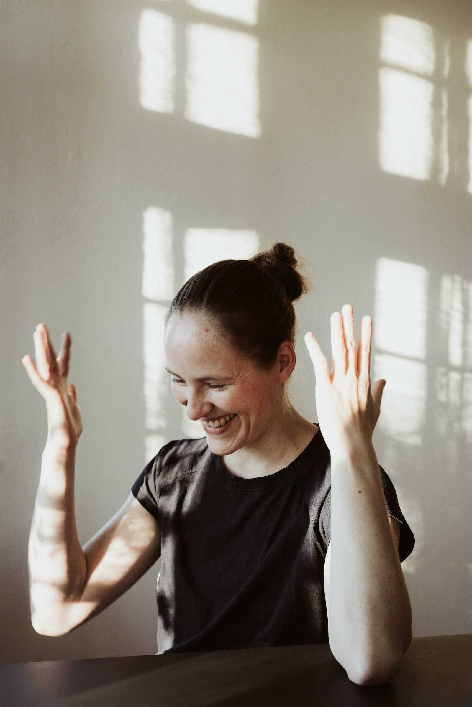 Lächelnde Frau mit hochgesteckten Haaren, die an einem Tisch sitzt, bei Sonnenlicht, das durch Fenster auf eine Wand scheint.