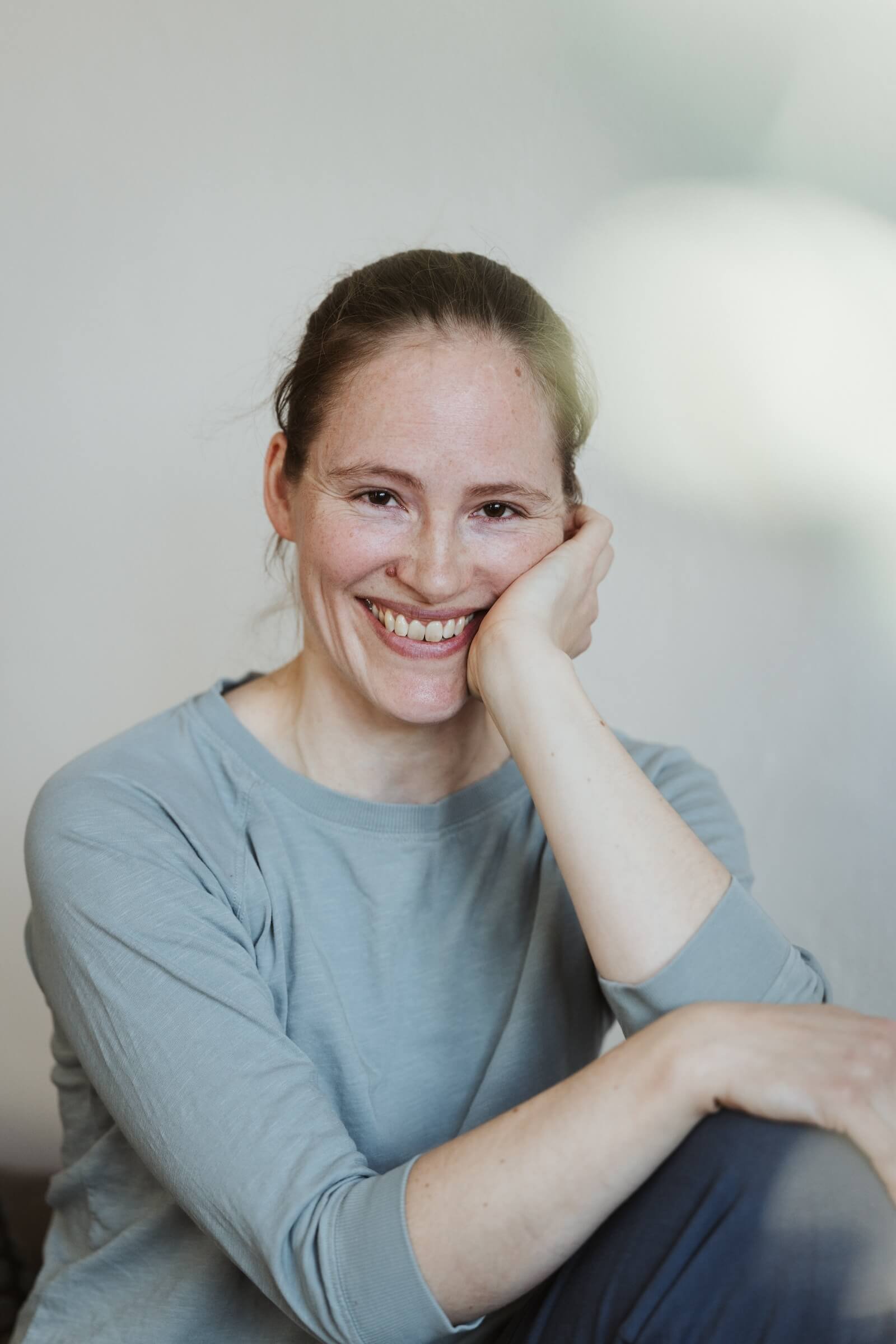 Eine lachende Frau mit braunen Haaren und Sommersprossen sitzt entspannt und lächelt. Sie trägt ein graues Oberteil und ruht ihren Kopf auf ihrer rechten Hand.