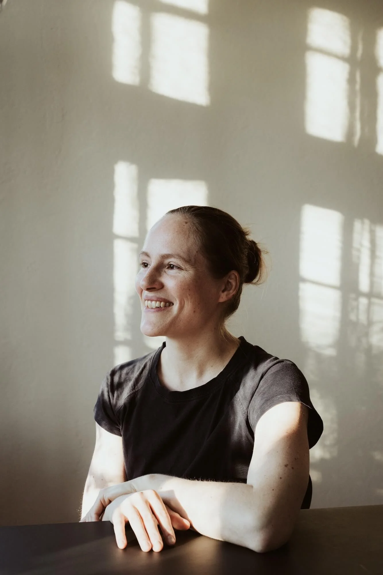 Lächelnde Frau mit dunkelhaarigem Zopf und schwarzen T-Shirt, sitzend an einem Tisch, Sonnenlicht reflektiert vom Fenster auf die Wand hinter ihr.