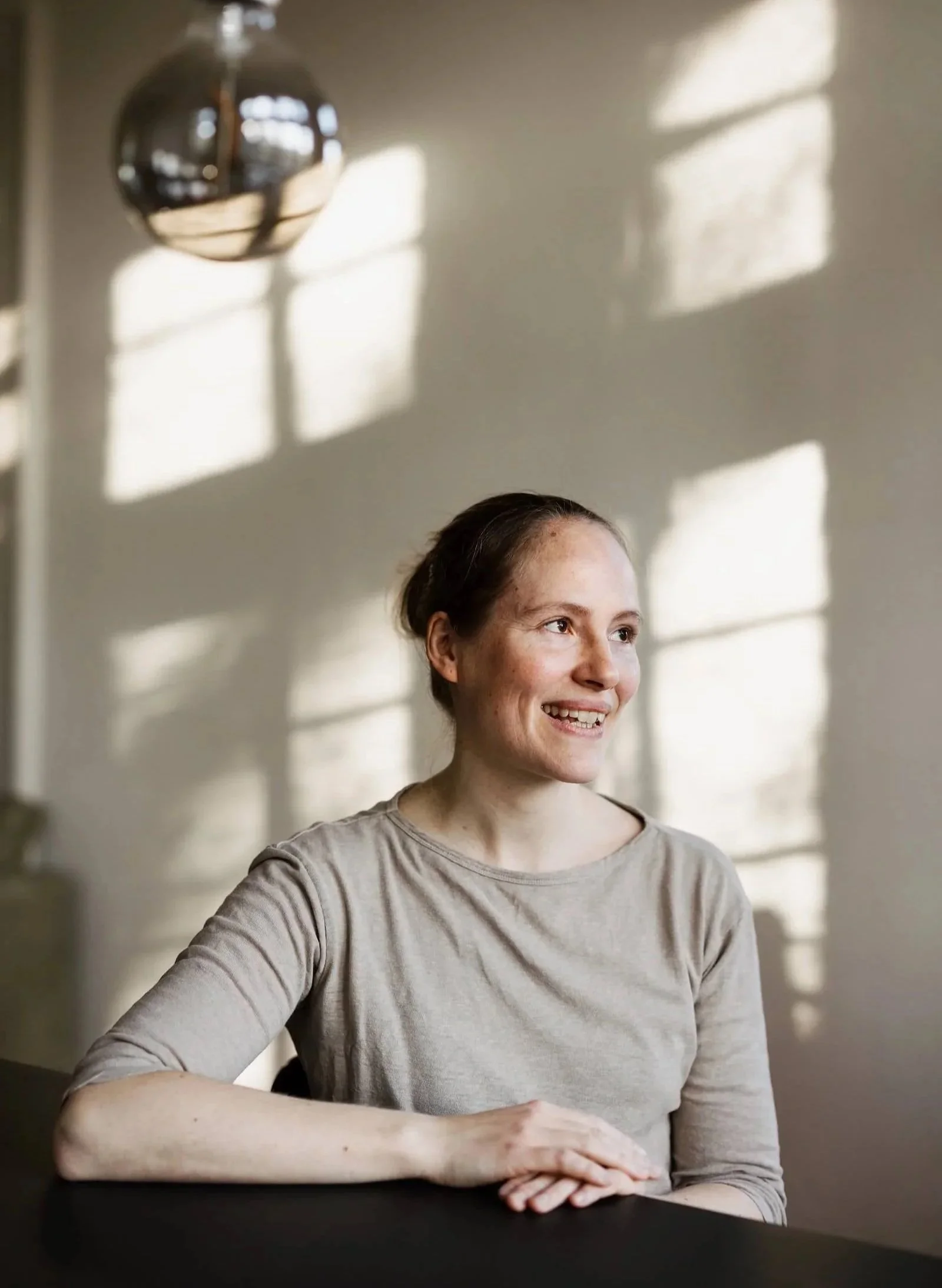 Eine lächelnde Frau sitzt an einem Tisch, im Hintergrund sind Schatten eines Fensters und eine Lampe sichtbar.