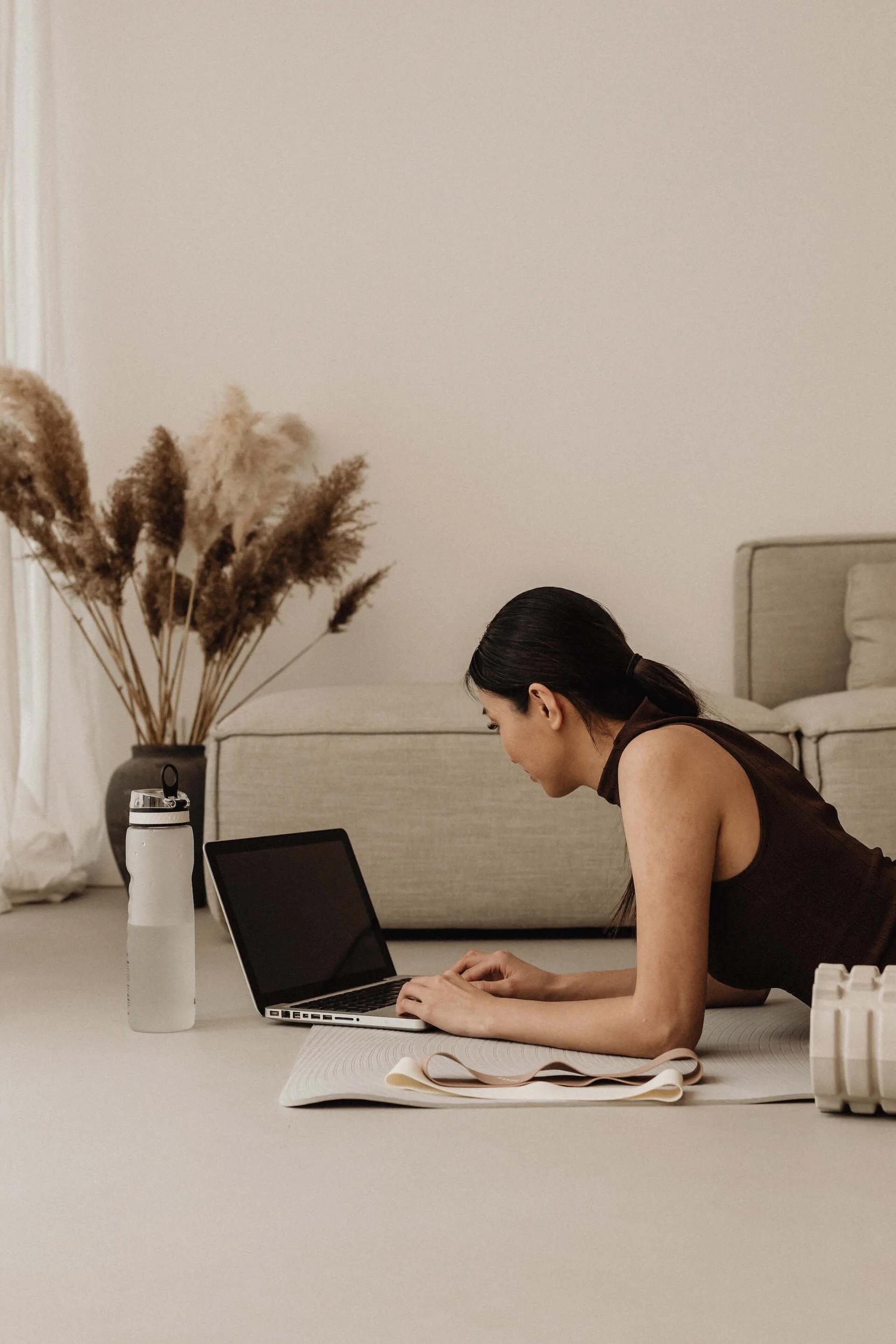 Frau trainiert Yoga auf dem Boden, arbeitet am Laptop, hat eine Wasserflasche und eine Yogamatte im Wohnzimmer mit einem Sofa, einer Vase mit Trockenblumen und einem Rollladen im Hintergrund.