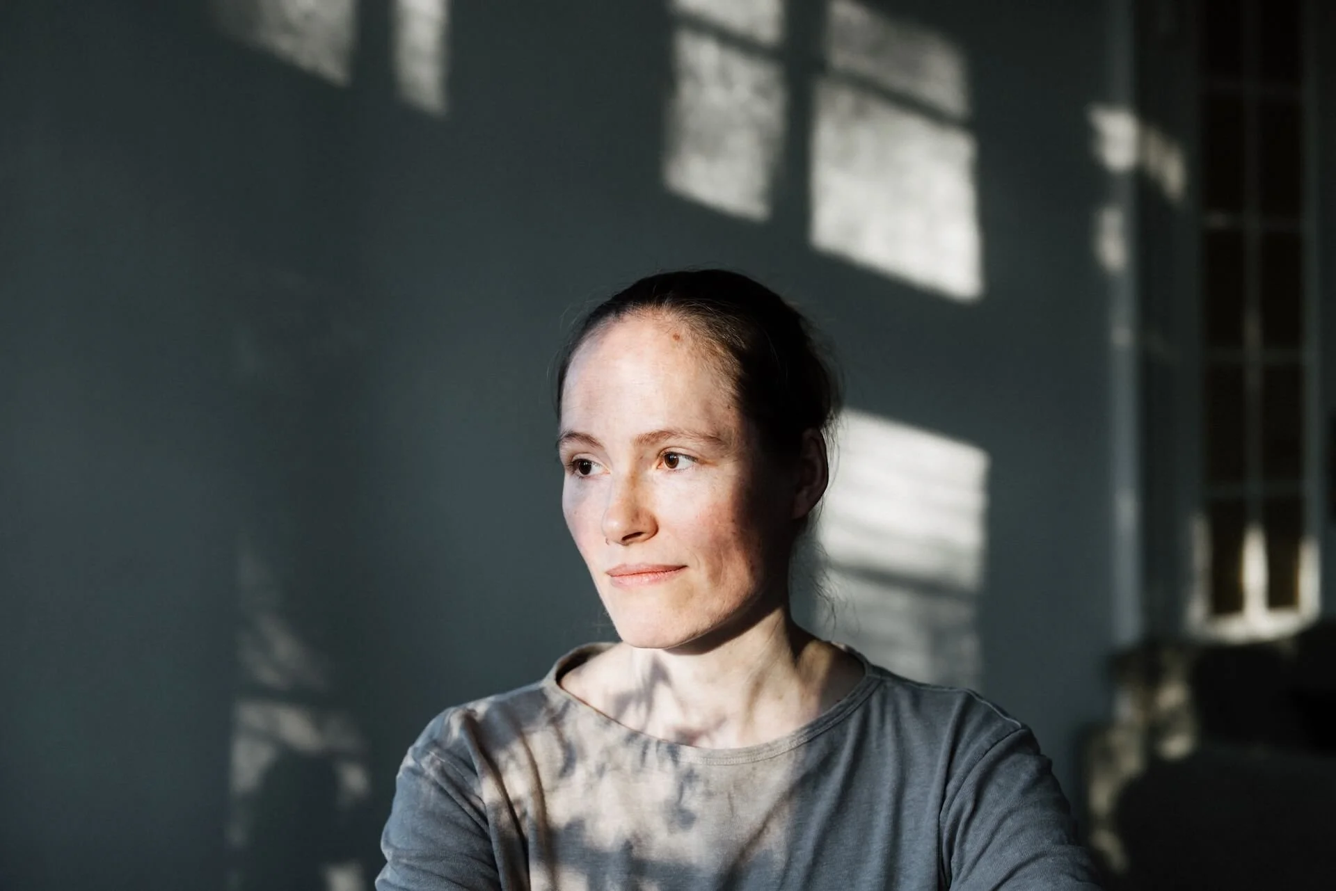 Nahaufnahme eines jungen Mädchens in einem grauen T-Shirt, das im Sonnenlicht an einer Wand sitzt, mit Schatten von Fenstern im Hintergrund.