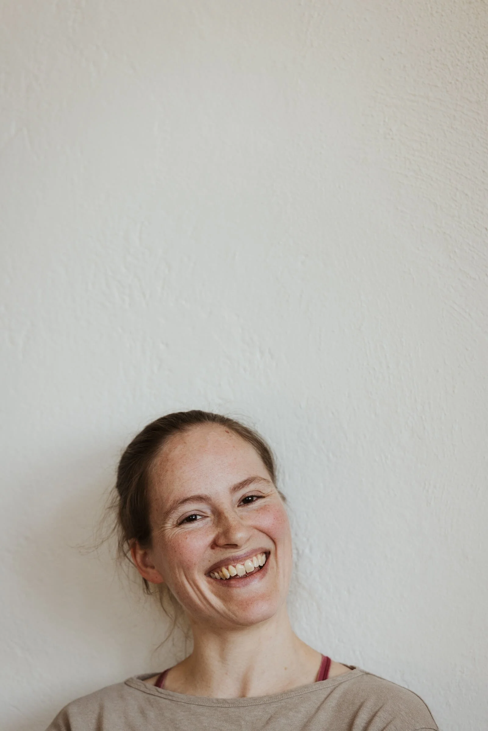Eine lachende Frau mit braunem Haar und Sommersprossen, die ein graues T-Shirt trägt, vor einer weißen Wand