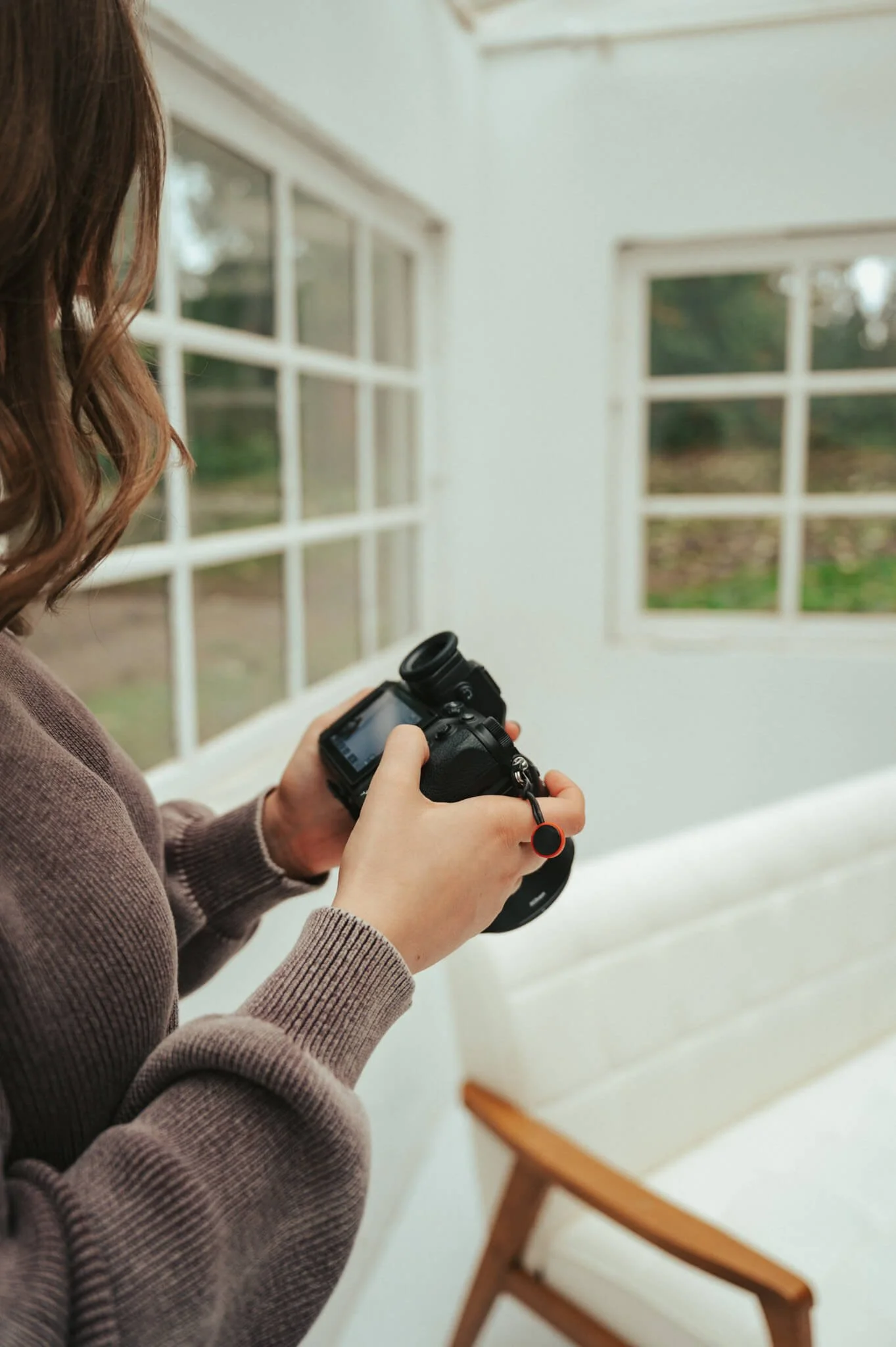 Person holding a camera indoors near large windows with a view of greenery outside.