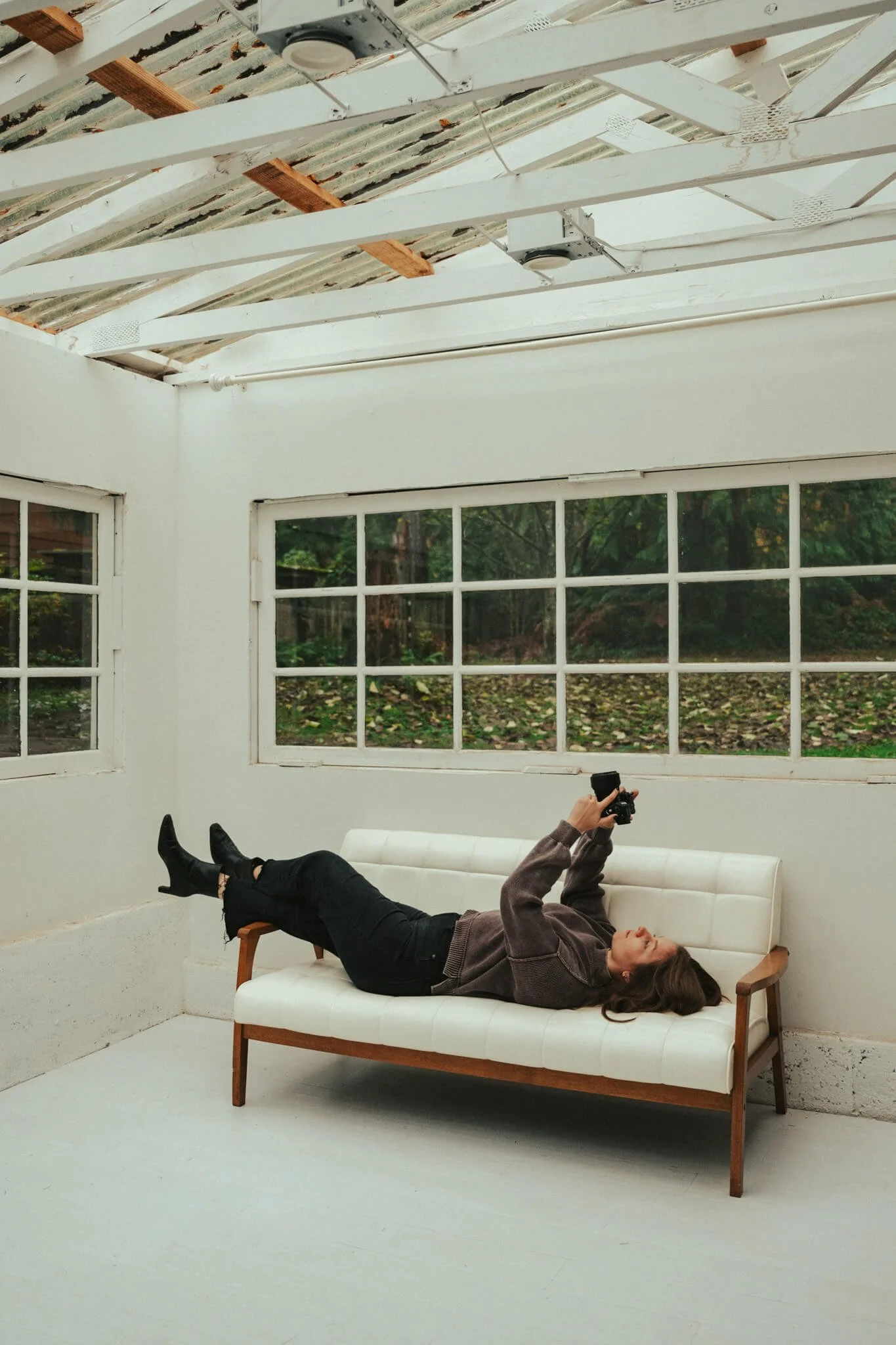 Woman lying on a white couch, holding a camera, inside a bright room with large windows and a partially exposed ceiling.