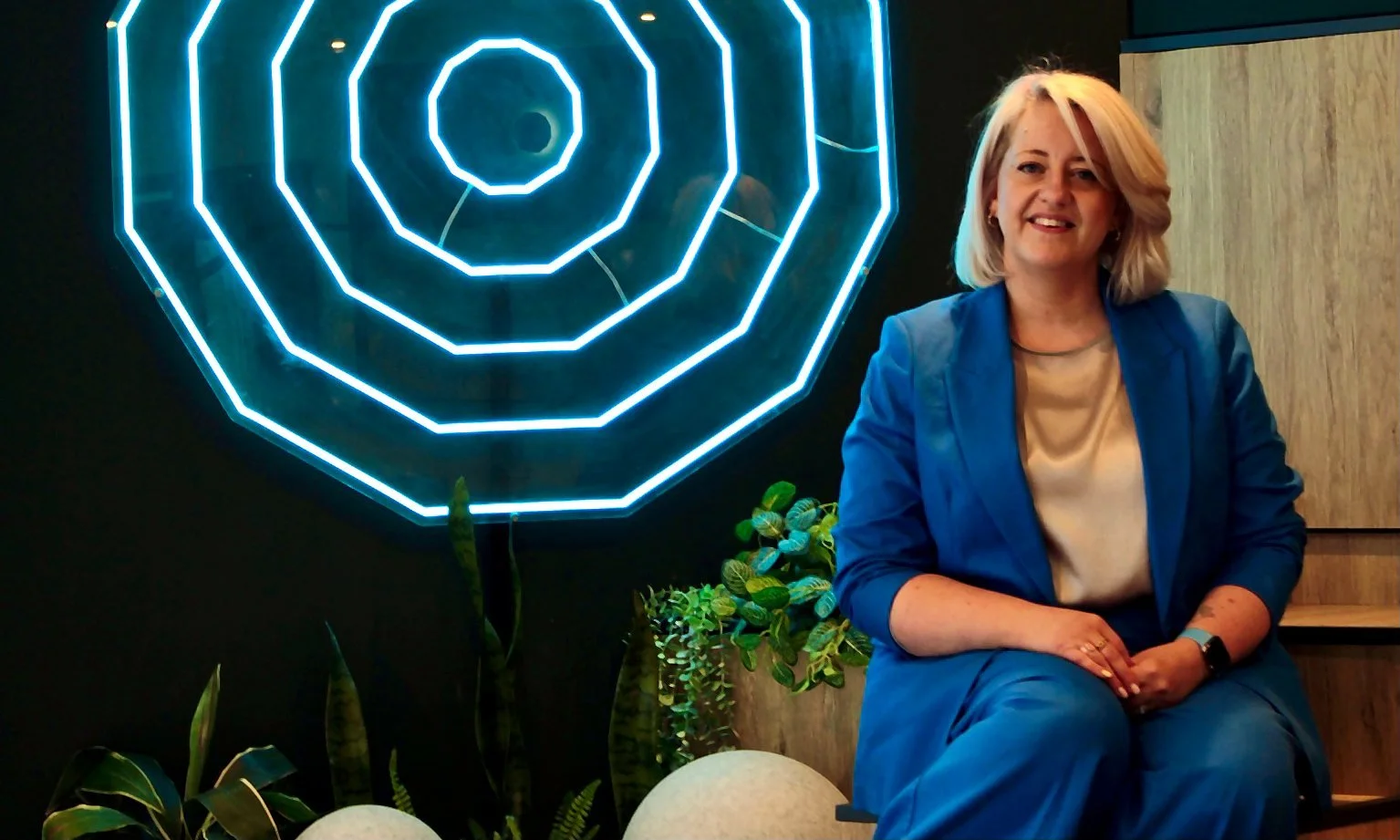 Jen in a blue suit sits beside a glowing geometric wall feature in a modern indoor setting