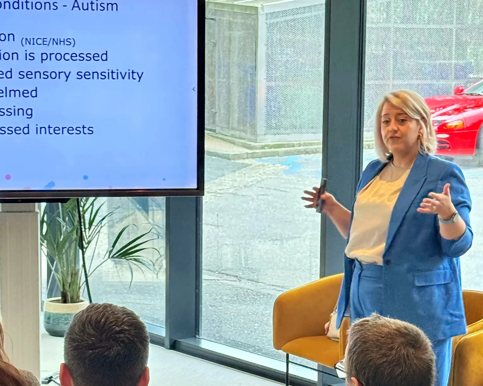 Jen delivers a presentation about autism to an audience, standing beside a large screen in a bright meeting space