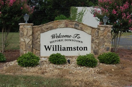 Welcome to Historic Downtown Williamston sign with stone border, surrounded by bushes and flowering trees.