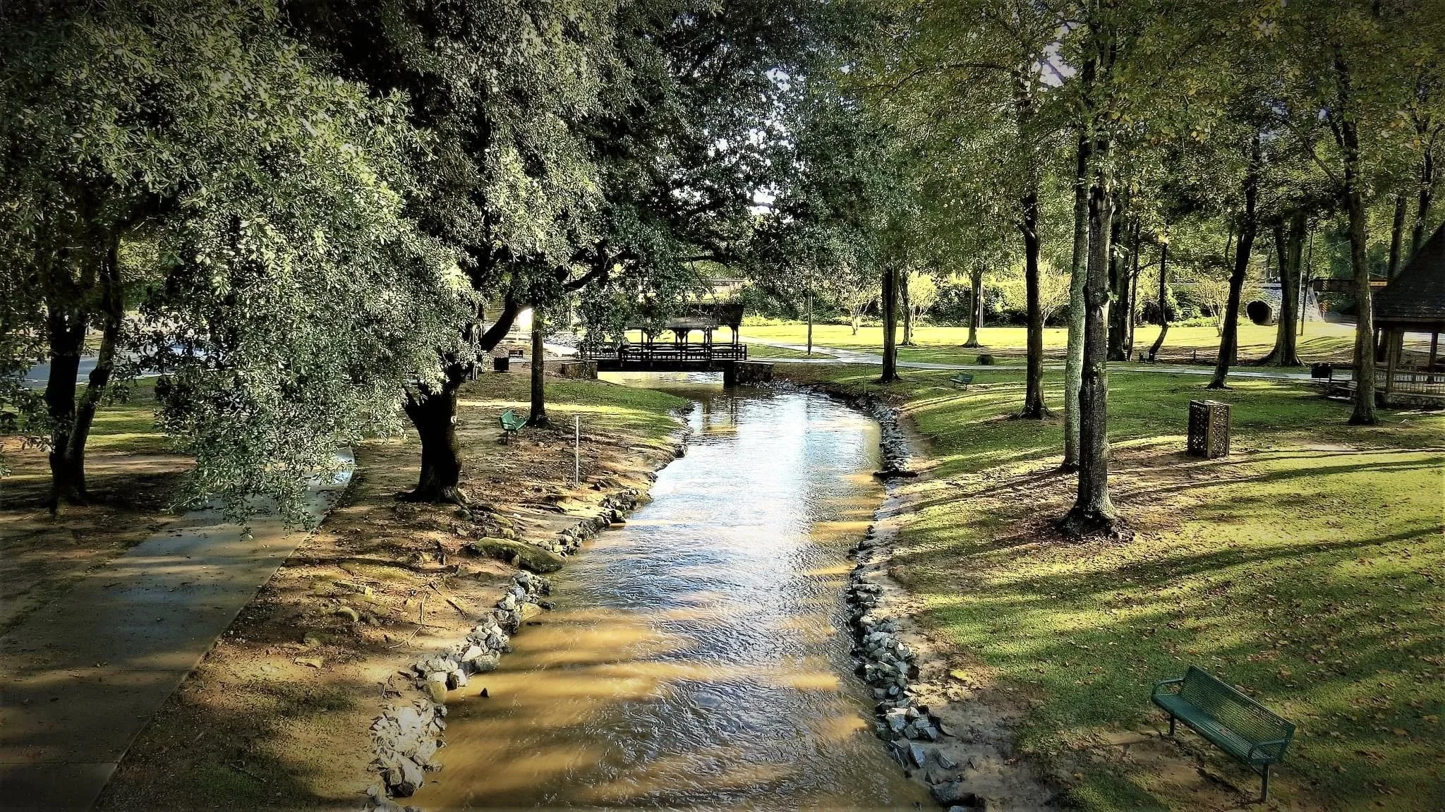 A peaceful park scene with a small stream running through, surrounded by trees and grassy areas, with walkways, benches, and a pavilion in the background.